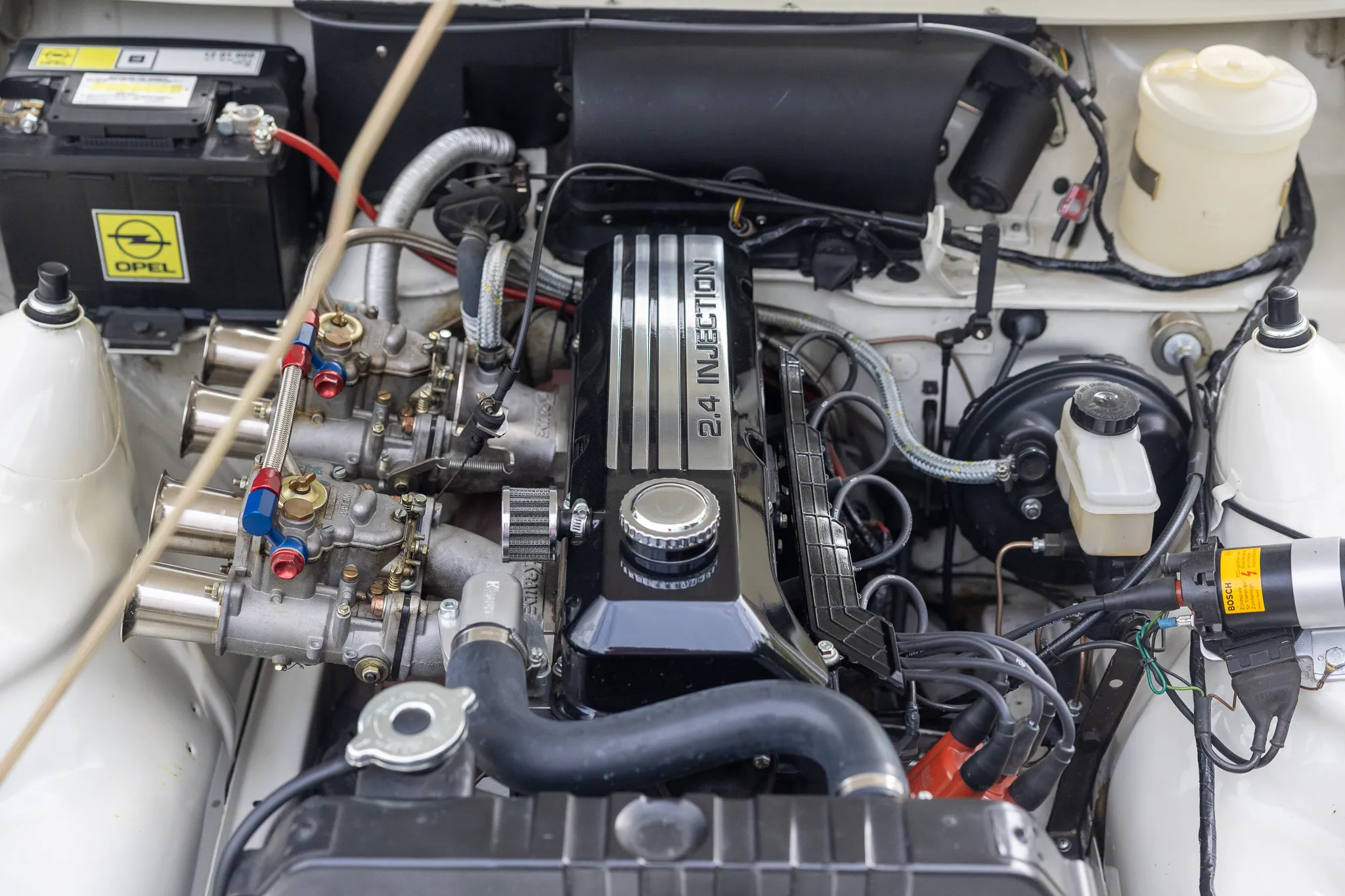 Motorraum eines Oldtimers mit dem Aufdruck „2.4 Injection“ auf dem Ventildeckel und dem Opel-Logo auf der Batterie.