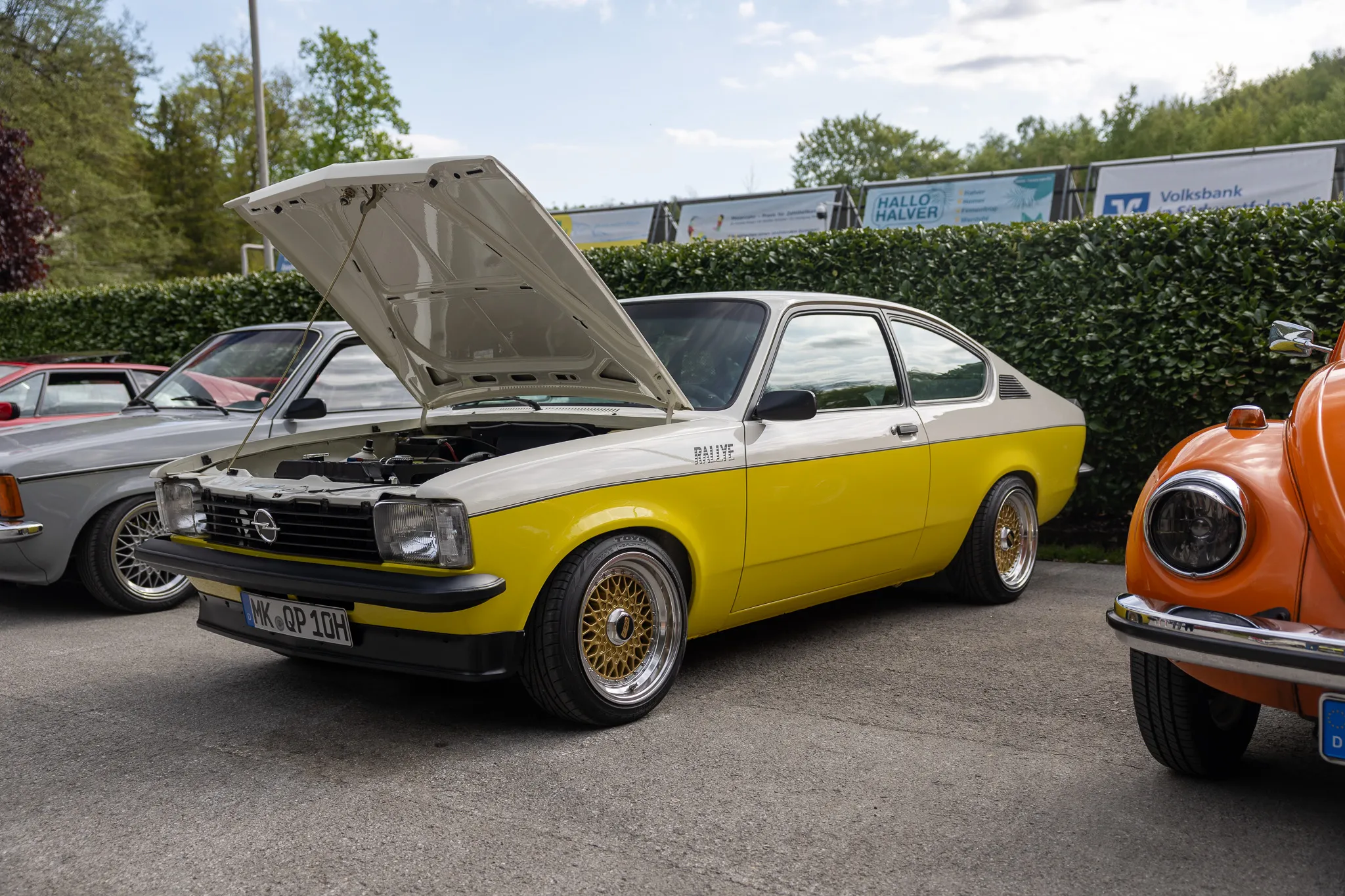 Gelber Opel Ascona mit geöffneter Motorhaube steht auf einem Oldtimertreffen neben einem orangefarbenen VW Käfer.