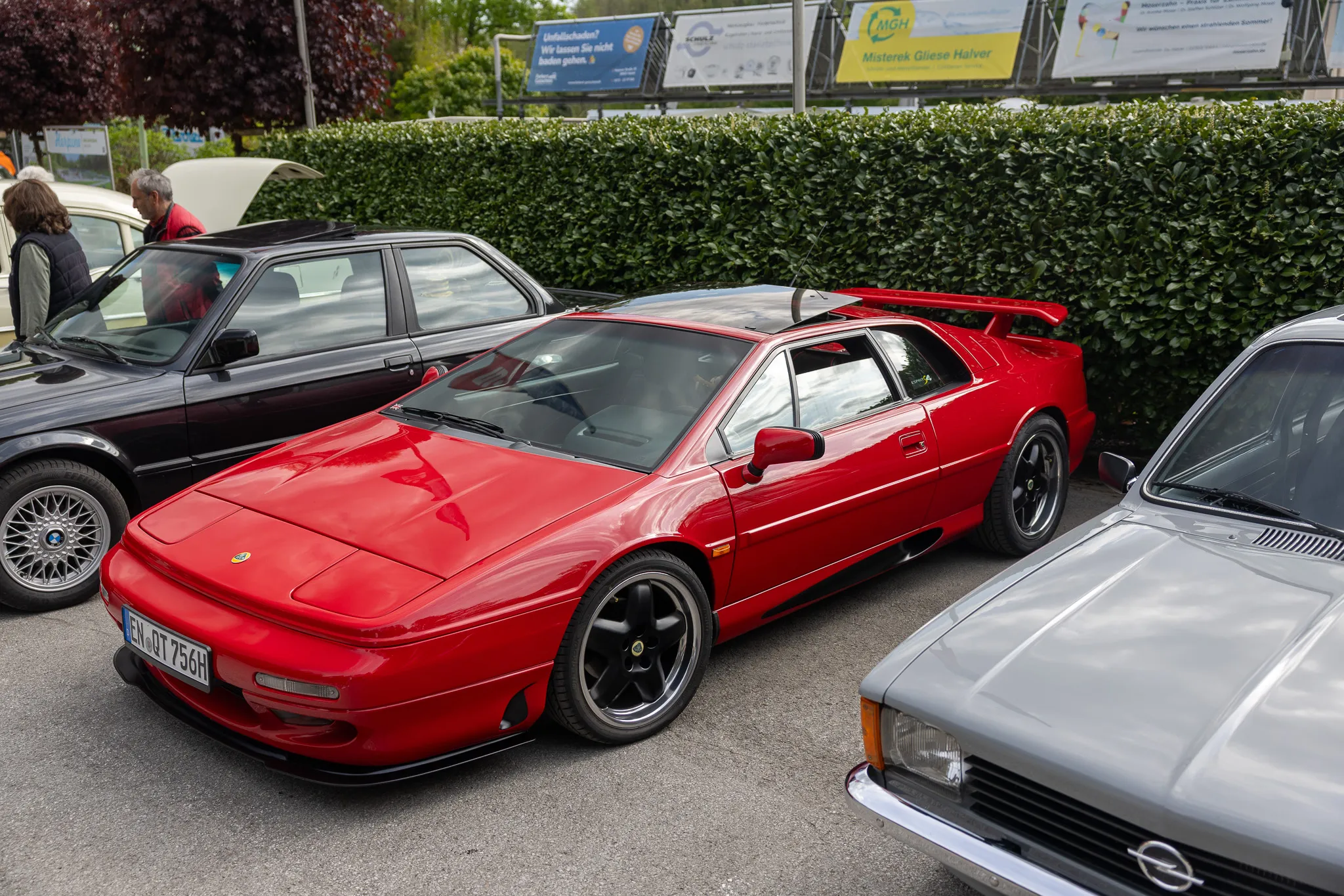 Roter Sportwagen Lotus steht im Mittelpunkt eines Oldtimertreffens, umgeben von weiteren Fahrzeugen und Zuschauern.