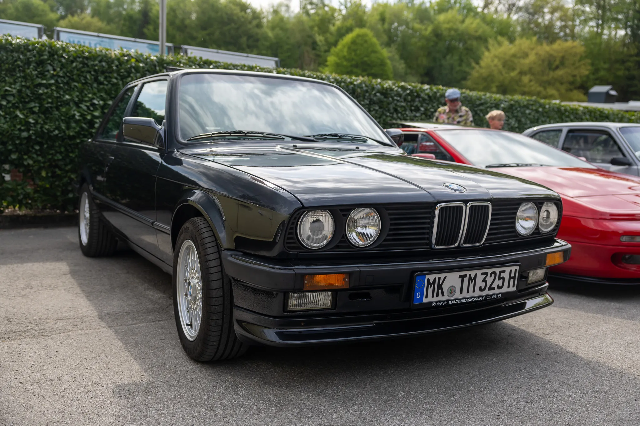 Schwarzer BMW E30 steht vor grüßen Hecken, Kennzeichen MK TM 325 H. Im Hintergrund ein rotes Auto und eine Person.