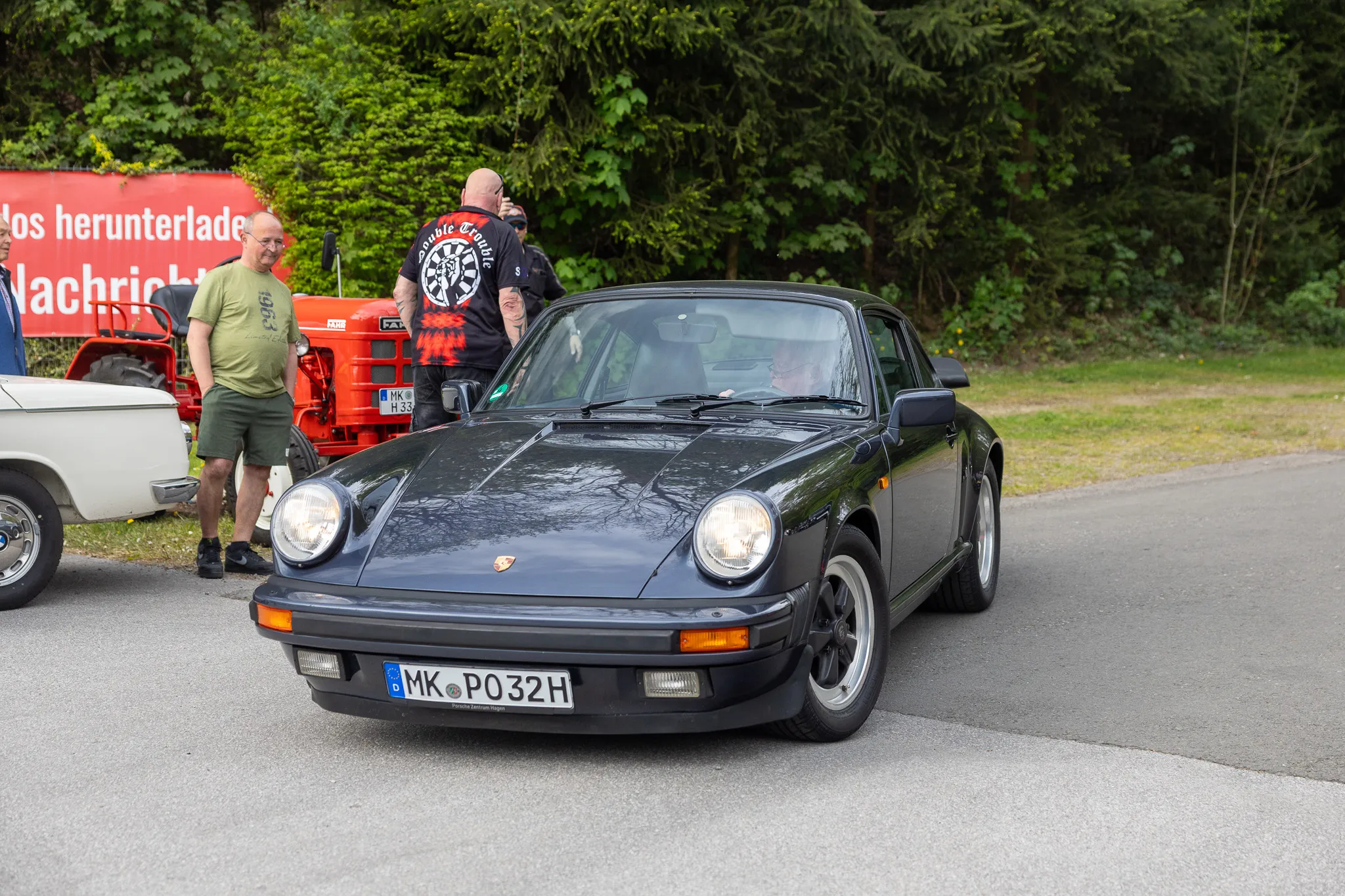 Dunkelblauer Porsche 911 steht auf einer Straße, während ein Mann in der Nähe des Fahrzeugs steht und ein weiterer Mann neben einem roten Traktor. Im Hintergrund Bäume.