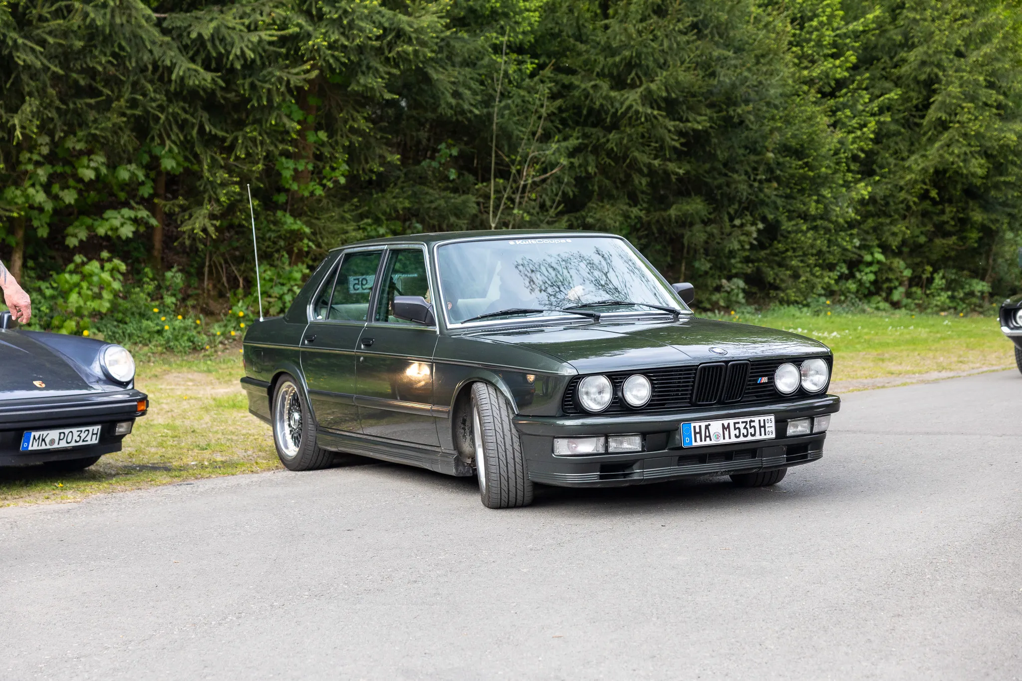 Dunkelgrüner BMW E30 steht auf einem Parkplatz vor Bäumen, ein weiteres Oldtimerauto ist links im Bild zu sehen.