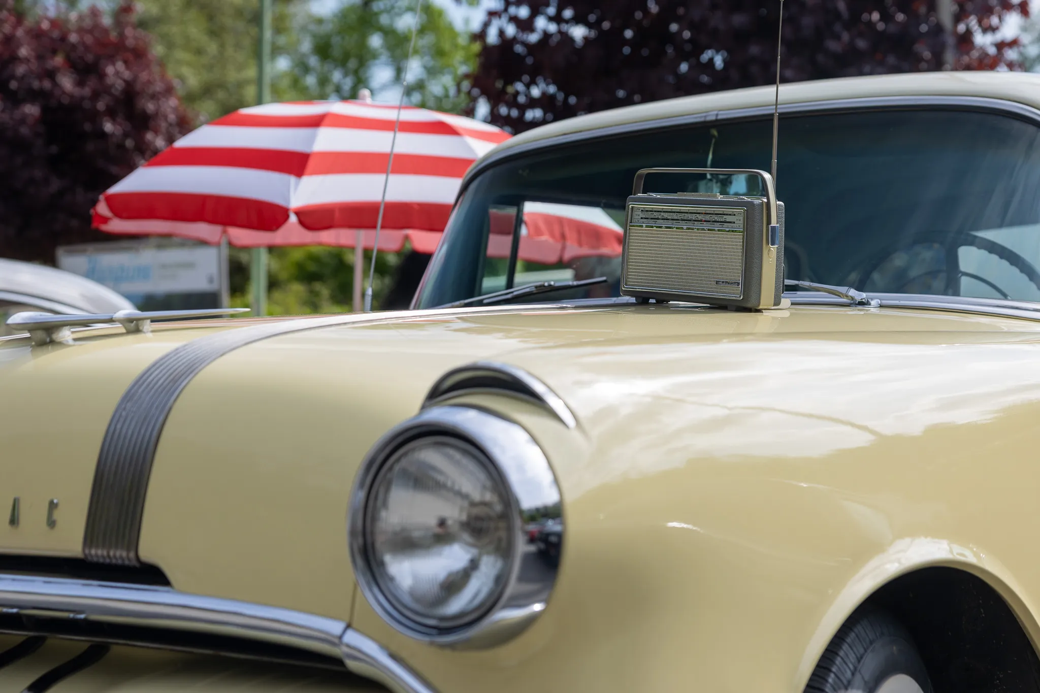 Beiger Oldtimer mit einem tragbaren Radio auf der Motorhaube, im Hintergrund ein rot-weiß gestreifter Sonnenschirm.