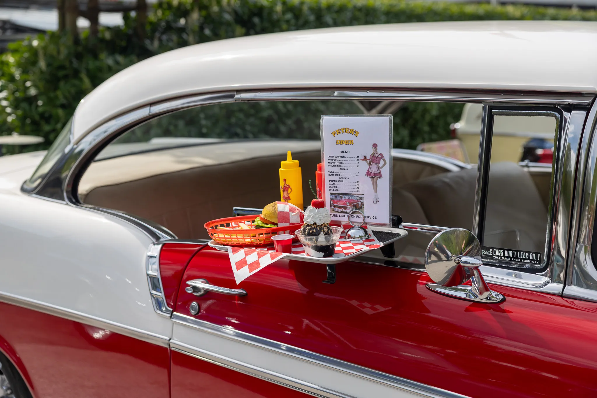 Rotes Oldtimerauto mit einem Tablett voller Fast Food darauf: Burger, Pommes, Getränke und eine Speisekarte eines Diners.