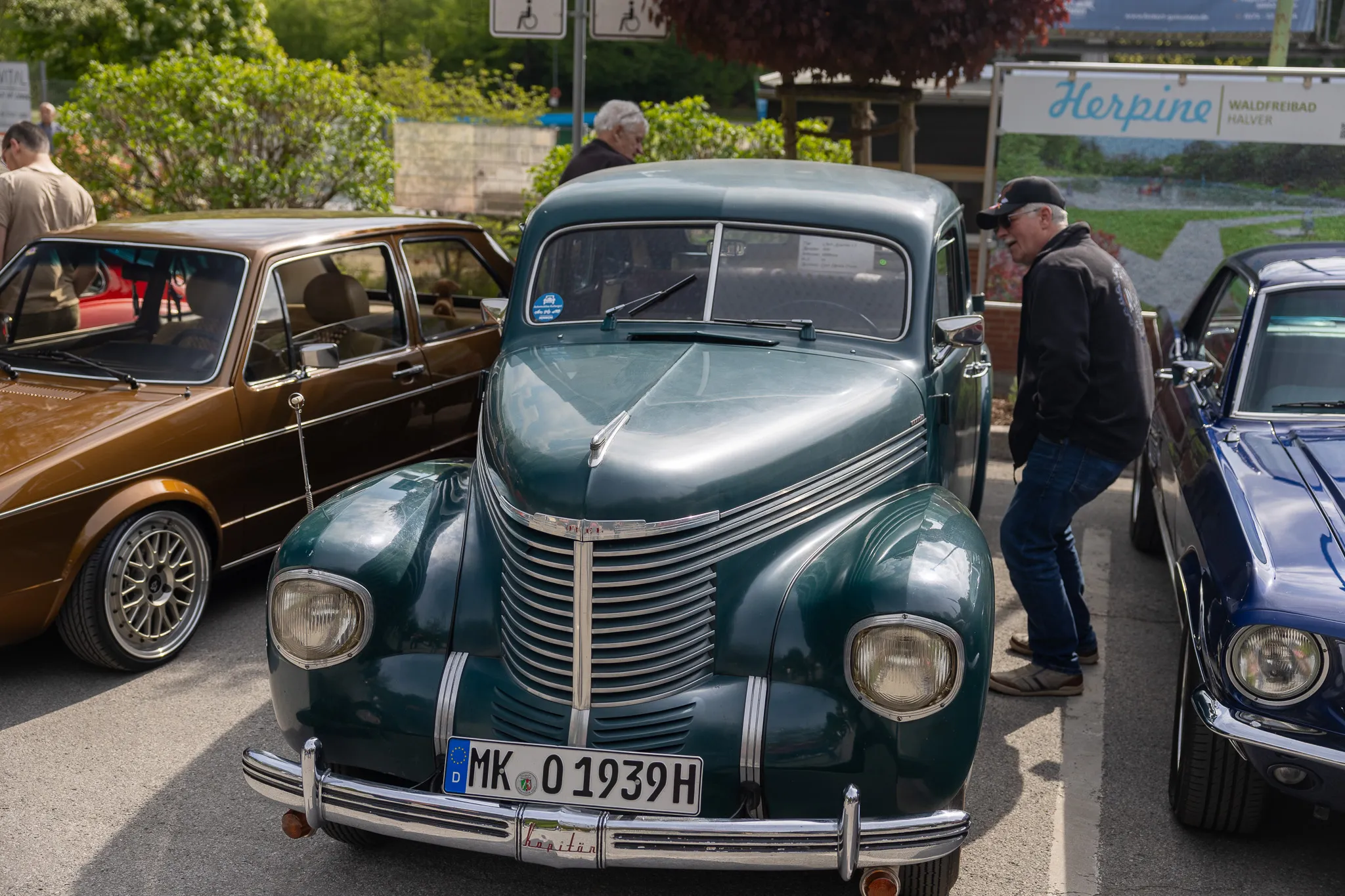 Ein dunkelgrüner Oldtimer mit Chromdetails steht im Mittelpunkt eines Oldtimertreffens, während ein Mann in der Nähe das Fahrzeug betrachtet.