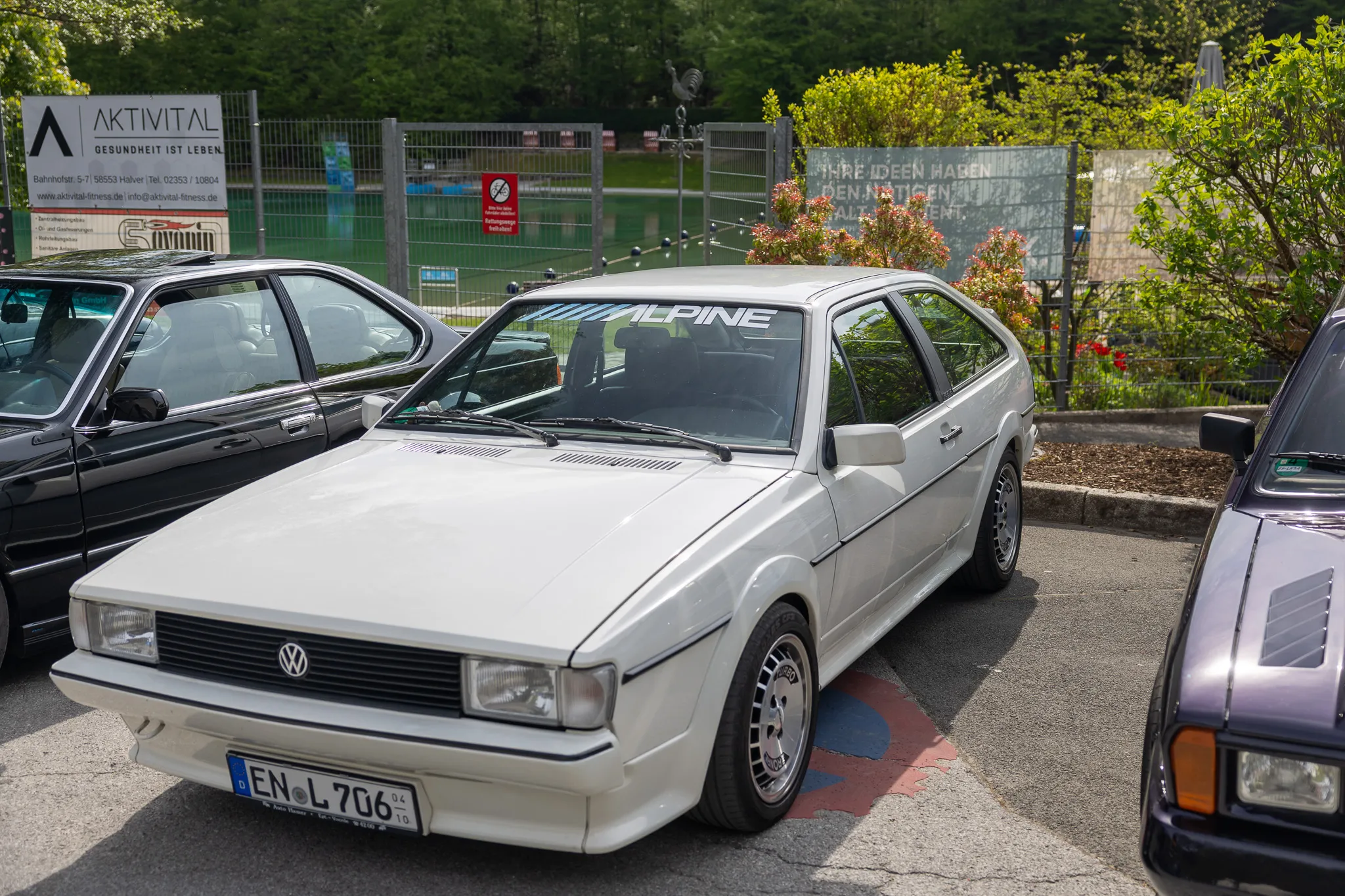 Weißer VW Scirocco mit dem Aufkleber „Alpine“ auf der Motorhaube steht vor einer grünen Hecke und weiteren geparkten Autos.