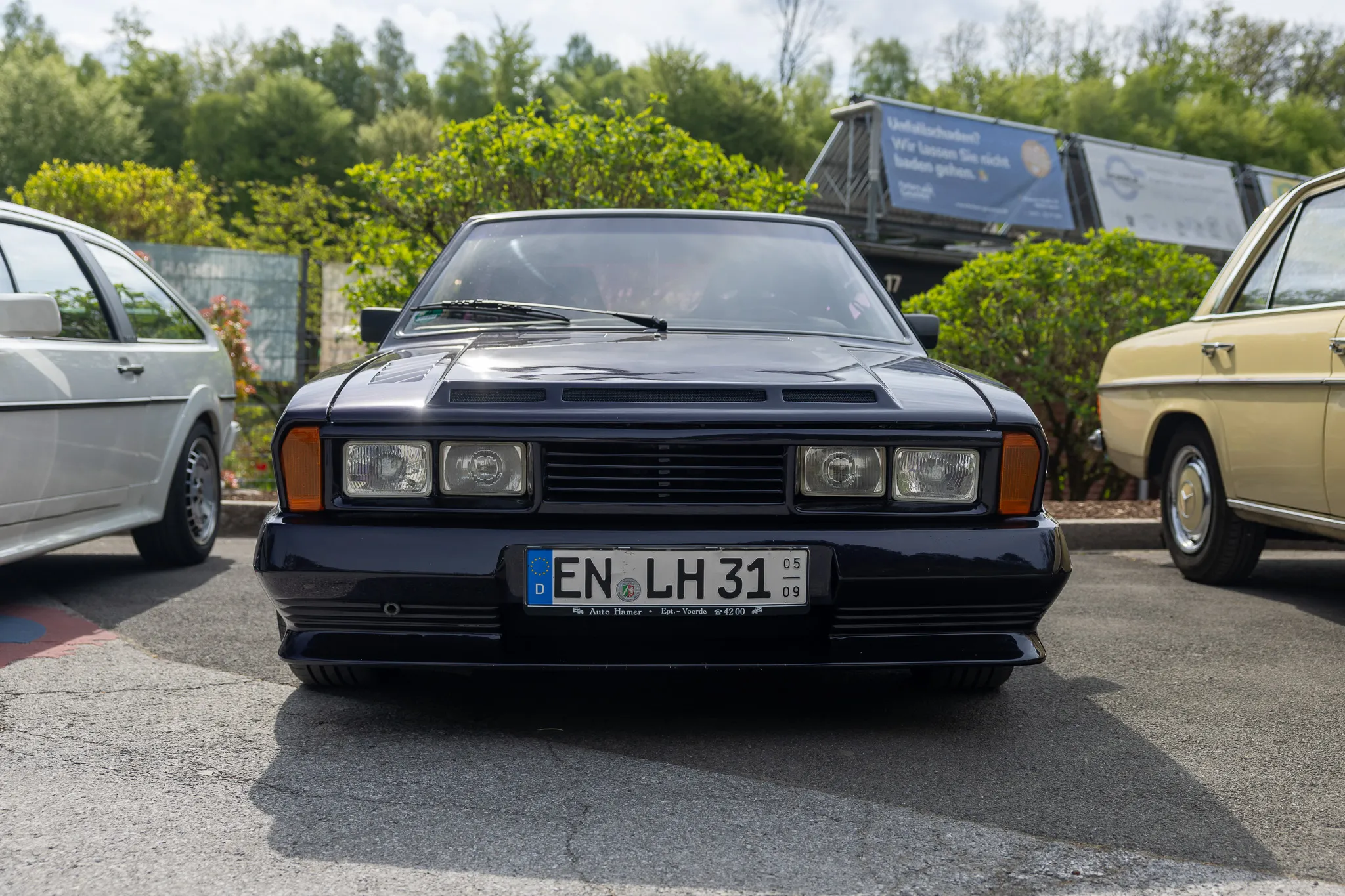 Dunkelblauer Volvo 240 mit deutschem Kennzeichen steht auf einem Parkplatz vor Bäumen und weiteren Fahrzeugen.
