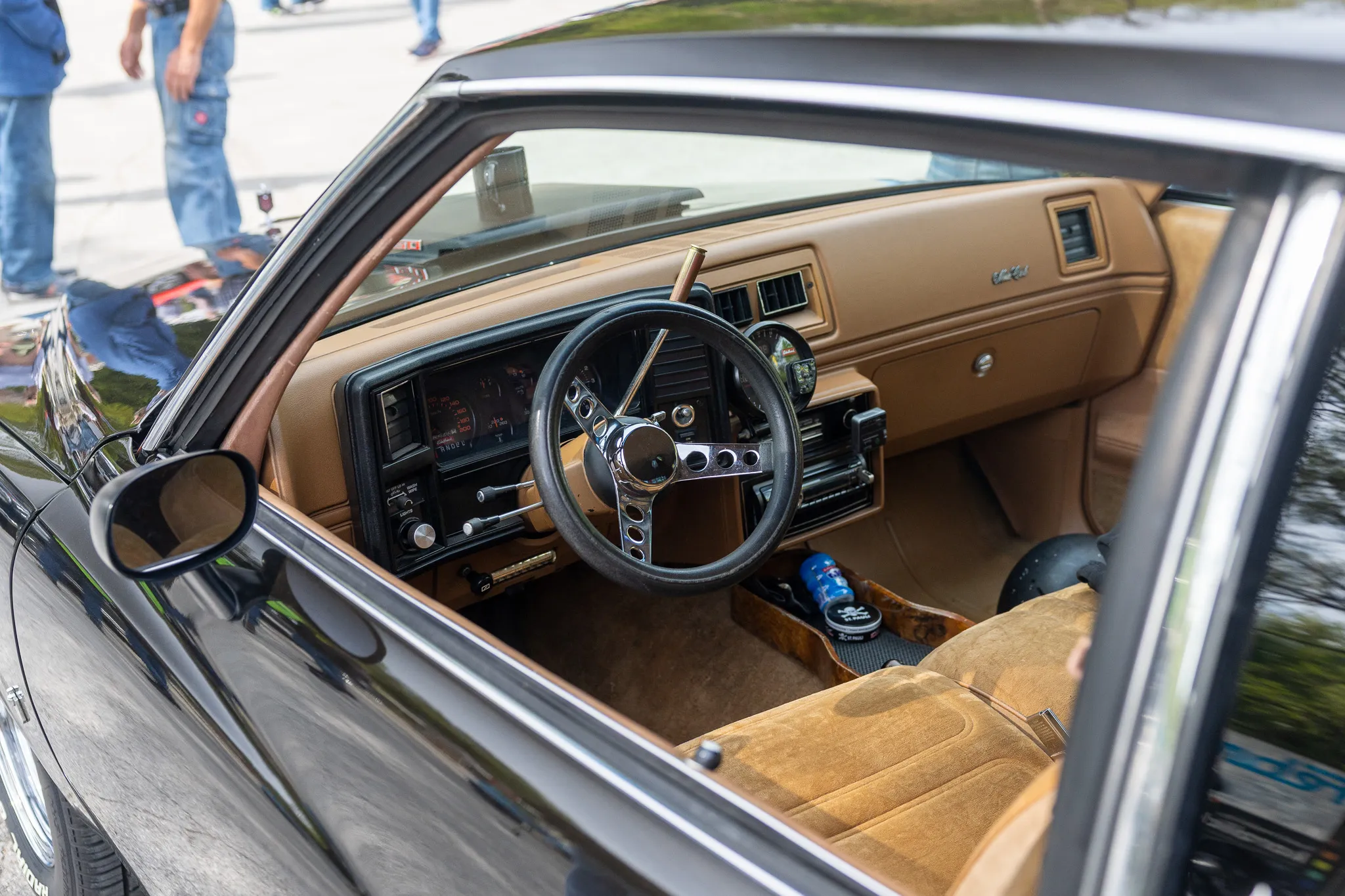 Innenraum eines klassischen Autos mit beigem Armaturenbrett, Lenkrad und Sitzen. Im Hintergrund sind Menschen zu sehen.