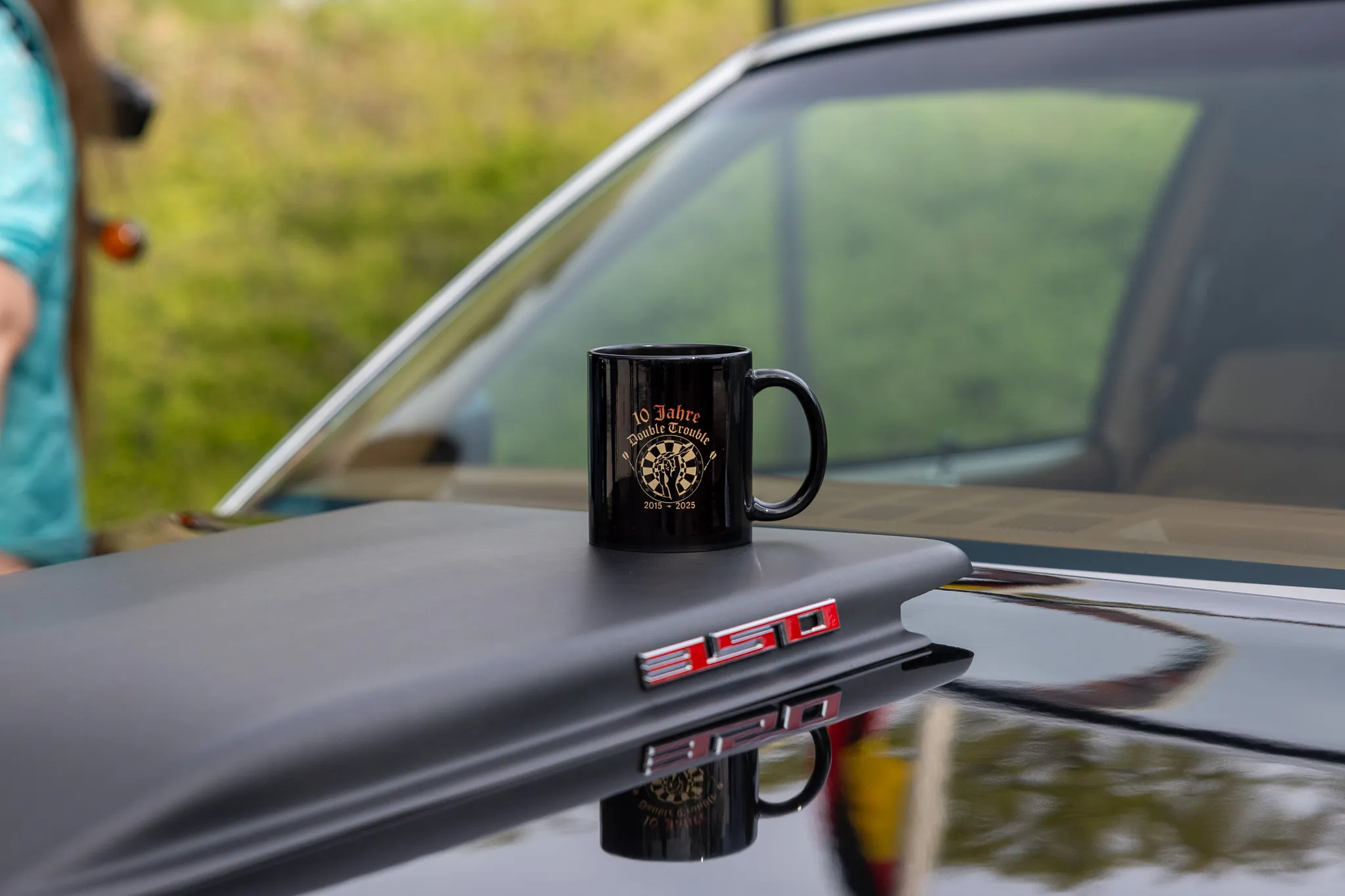 Schwarzer Kaffeebecher mit Aufdruck steht auf der Motorhaube eines Oldtimers. Im Hintergrund sind weitere Fahrzeuge und Bäume zu sehen.