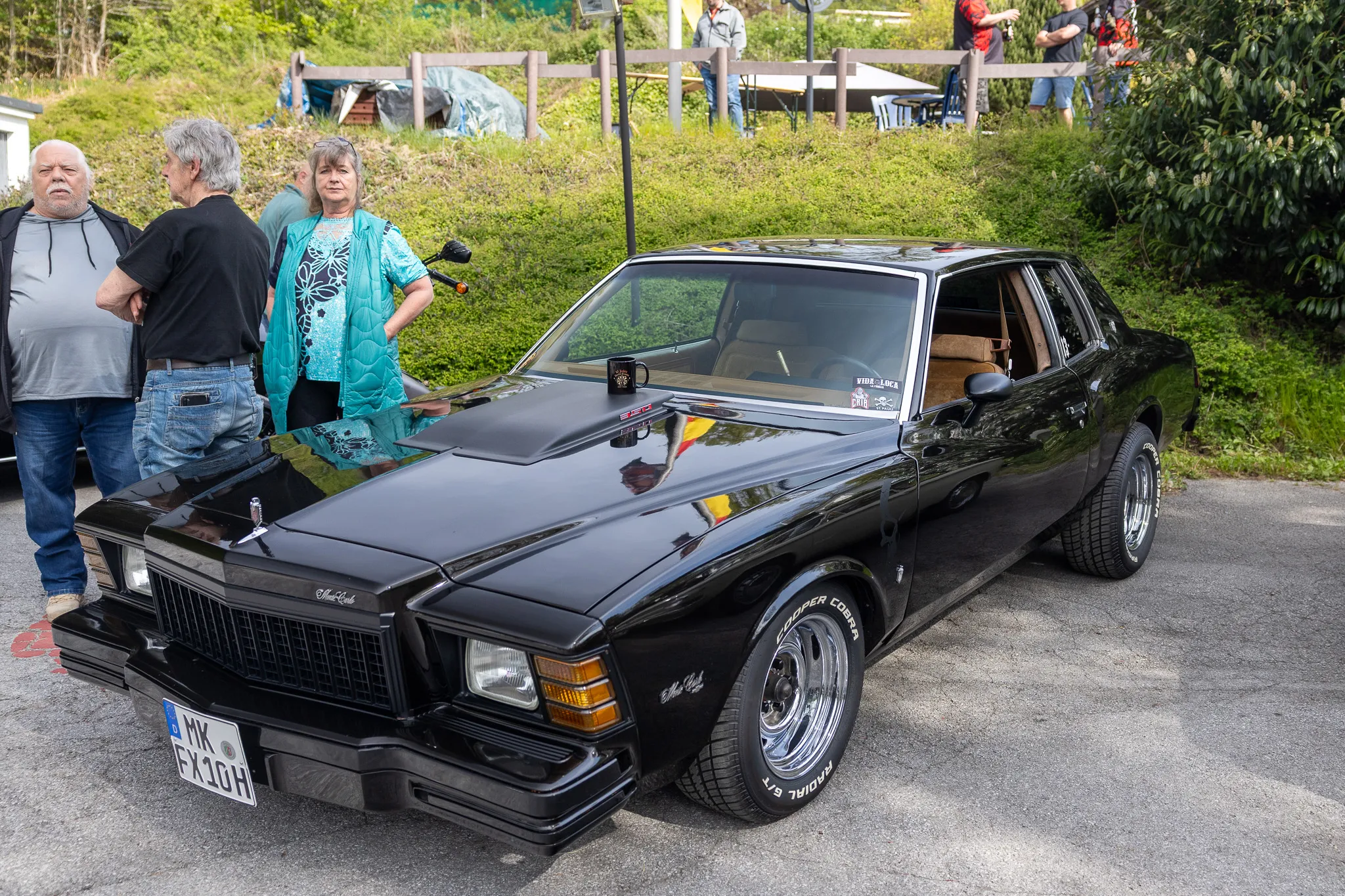 Schwarzer Oldtimer mit Chromdetails steht auf einem Parkplatz, umgeben von Zuschauern. Im Hintergrund sind weitere Personen und Zelte zu sehen.