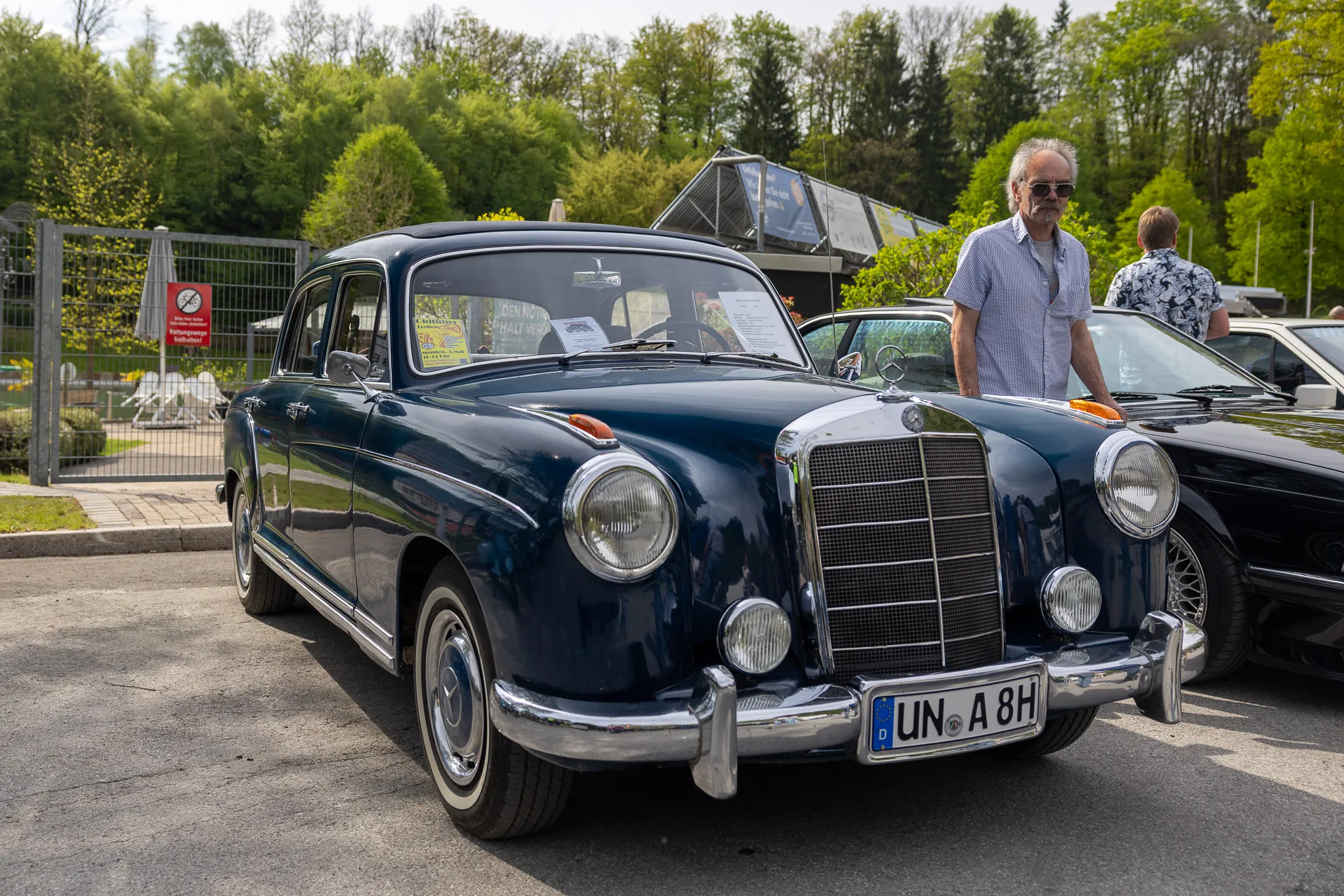 Dunkelblauer Mercedes-Oldtimer mit Chromdetails steht auf einem Parkplatz, ein Mann mit Hemd und Sonnenbrille steht daneben.