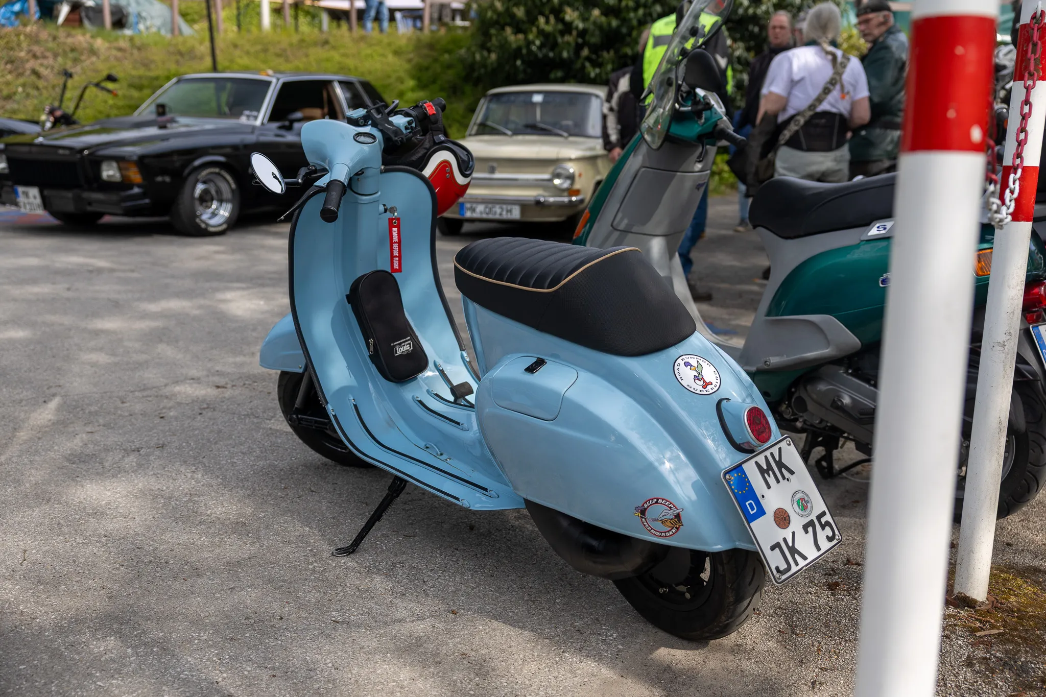 Hellblauer Vespa Roller mit Taschen und Aufklebern steht auf einem Parkplatz neben anderen Fahrzeugen und Zuschauern.