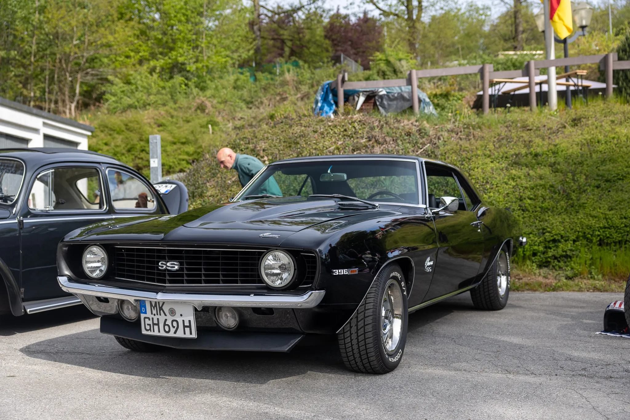 Schwarzer Chevrolet Camaro SS aus den frühen 1970er Jahren mit Chromdetails und dem Kennzeichen MK-GH 69 H steht auf einem Parkplatz vor Bäumen und Büschen.