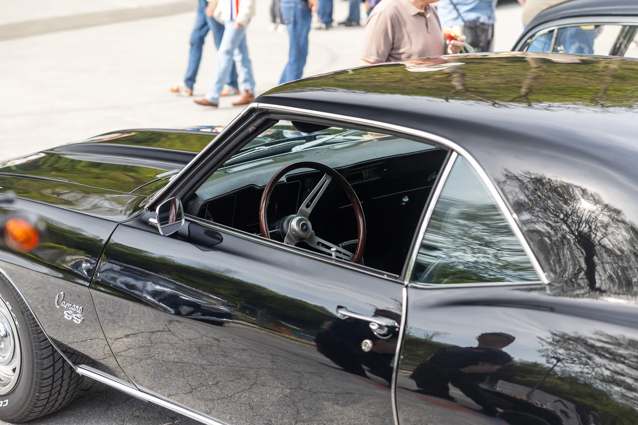 Schwarzer Oldtimer Camaro mit geöffneter Fahrertür, das Armaturenbrett und das Lenkrad sind sichtbar. Im Hintergrund sind Menschen auf einem Parkplatz zu erkennen.
