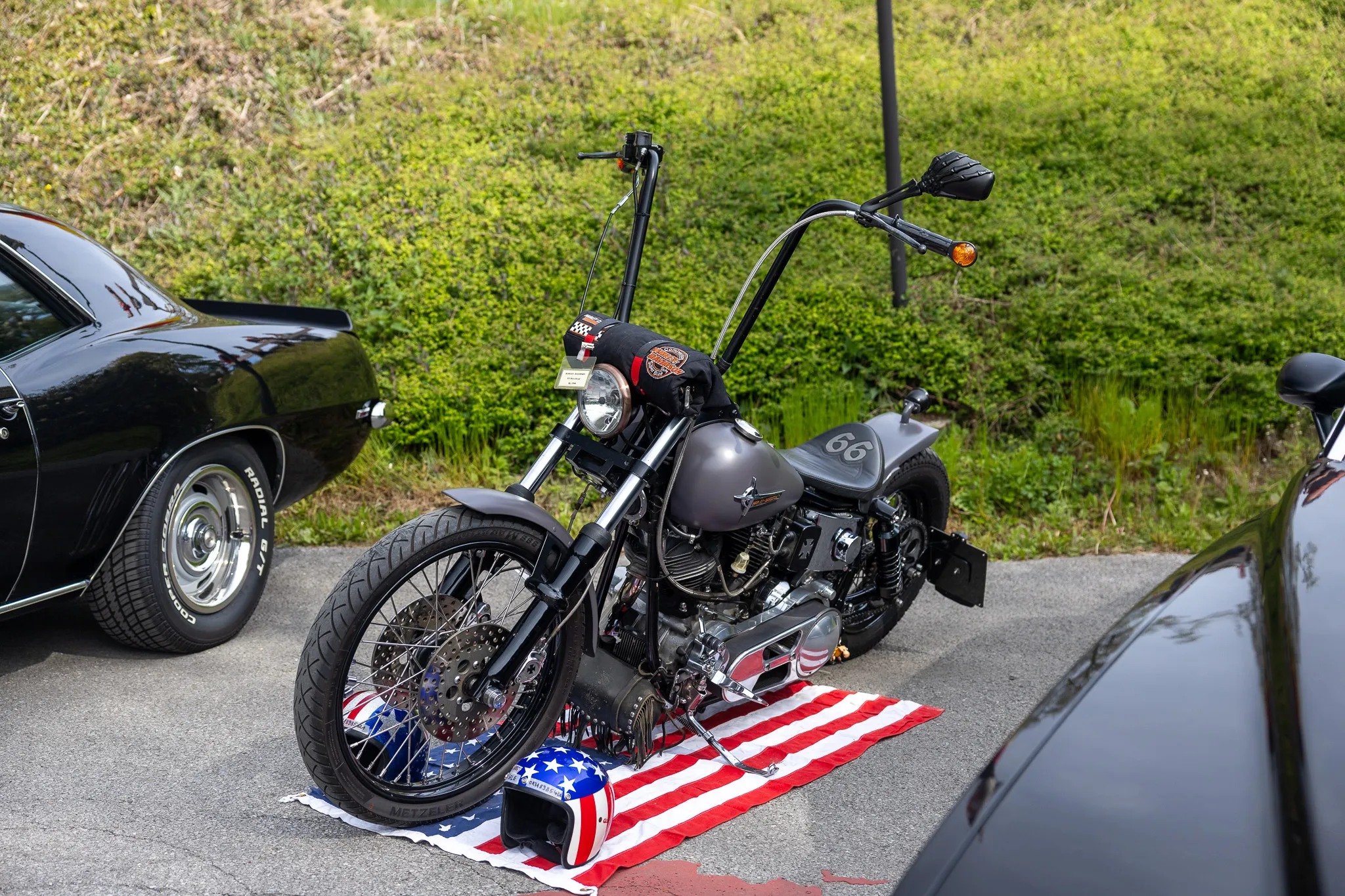 Dunkelgraues Chopper-Motorrad mit hohem Lenker steht auf einer amerikanisch gestalteten Matte vor geparkten Autos.