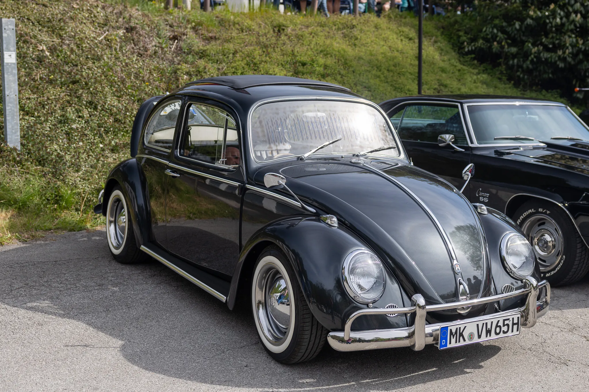 Schwarzer VW Käfer steht auf einem Parkplatz, daneben ein weiterer Oldtimer. Im Hintergrund sind Bäume und Menschen zu sehen.