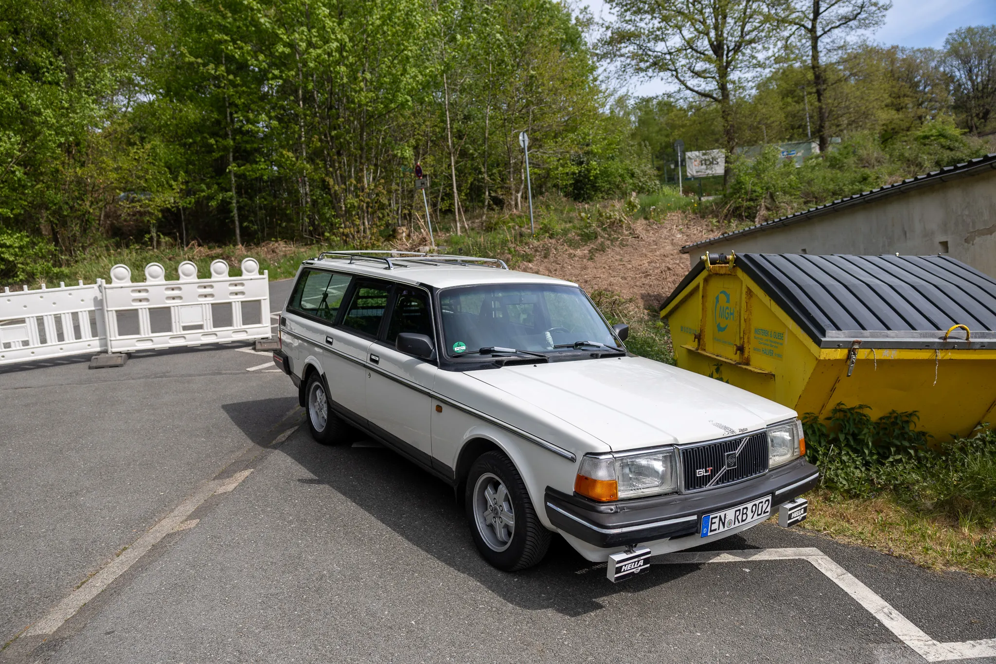 Weißer Volvo Kombi aus den 1980er Jahren steht auf einem Parkplatz vor einer Absperrung und einem gelben Container.