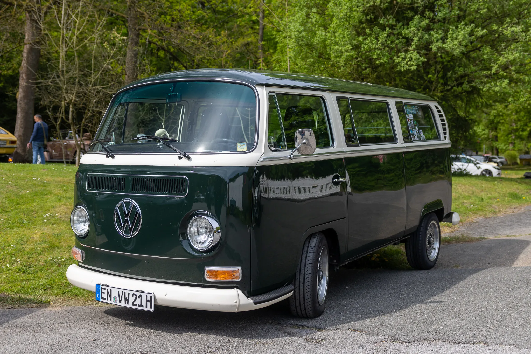 Grüner VW-Bus Typ 2 (T1) mit weißem Dach steht auf einem Parkplatz vor Bäumen. Das Kennzeichen ist teilweise verdeckt.