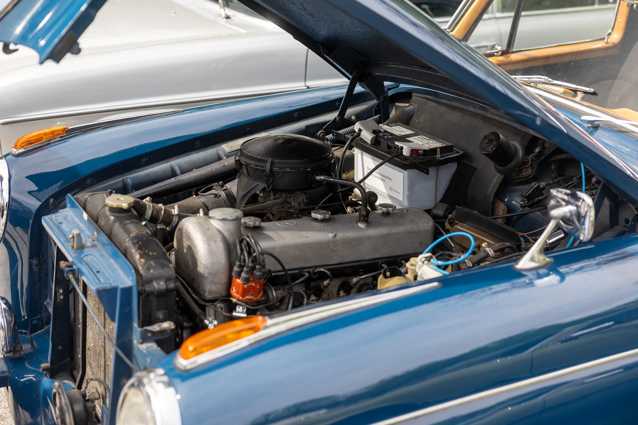 Geöffneter Motorraum eines klassischen dunkelblauen Autos mit einem weißen Batteriekasten und verschiedenen Metallteilen.