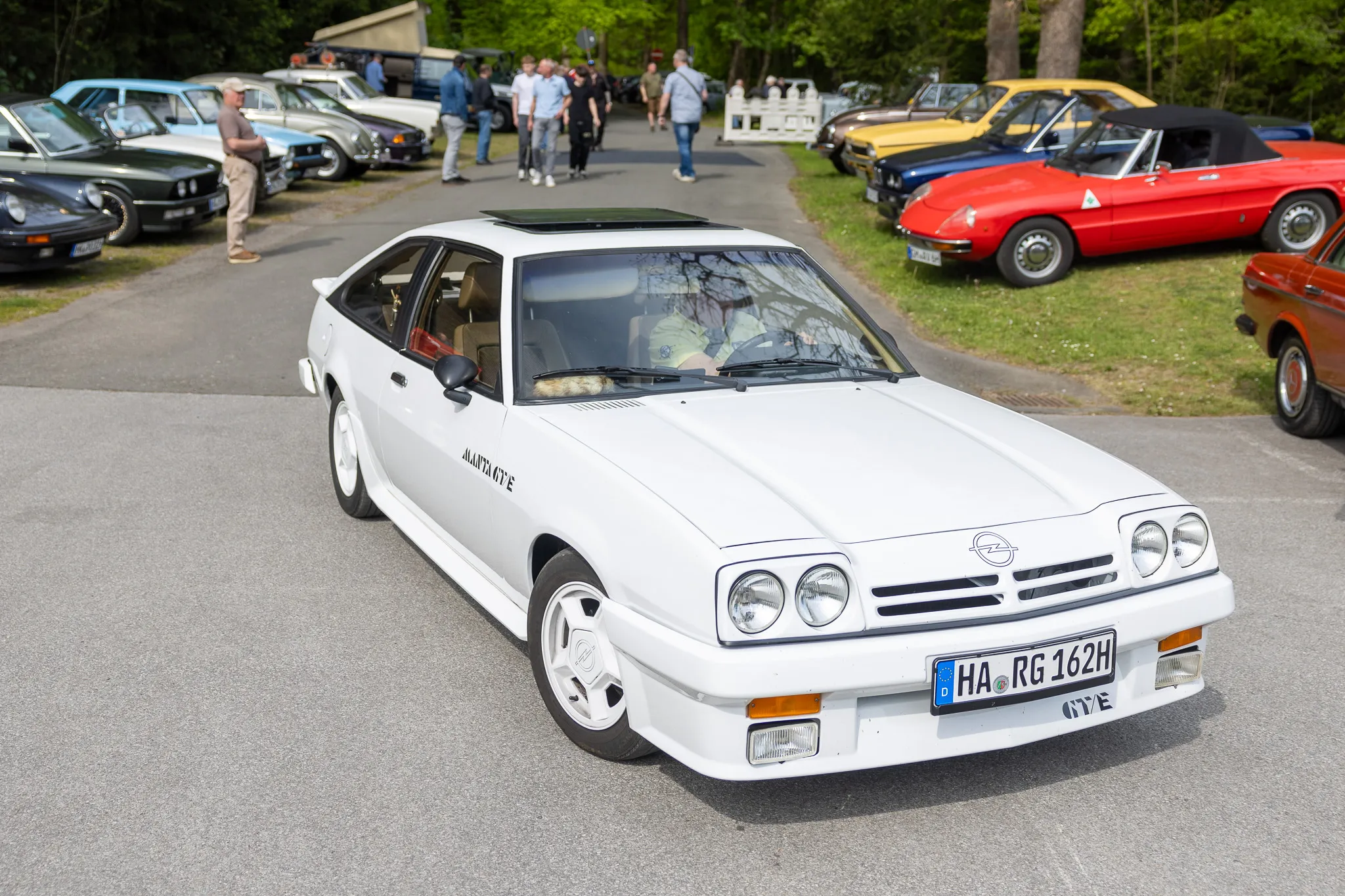 Weißer Opel Monza mit Kennzeichen HA-RG 126H steht auf einem Parkplatz vor einer Gruppe von Oldtimern und Besuchern.