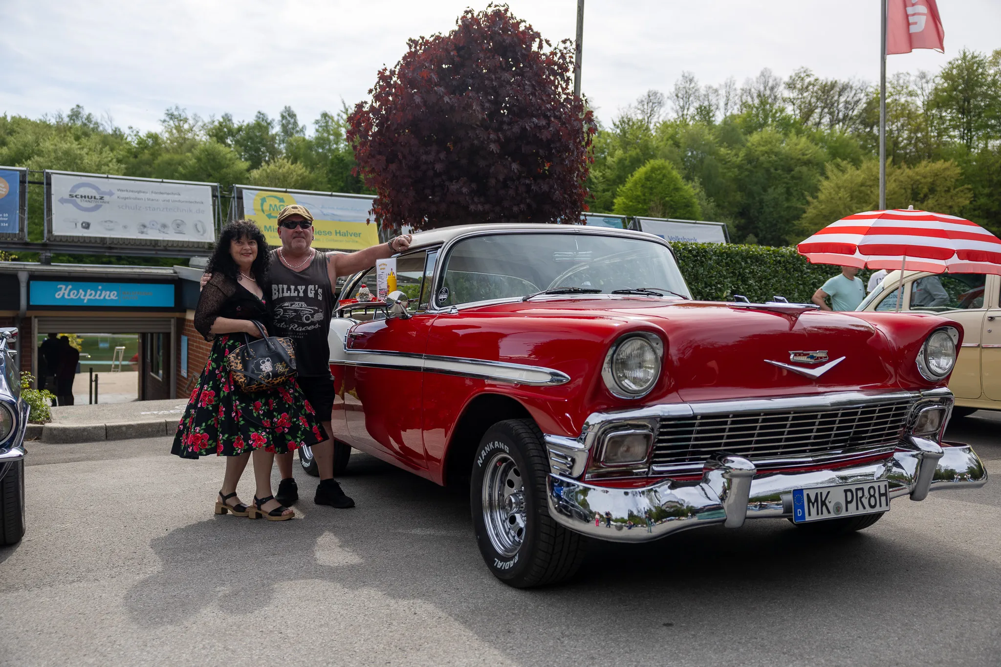 Ein Paar steht vor einem roten Oldtimer der 1950er Jahre auf einem Parkplatz, im Hintergrund ein Gebäude und weitere Fahrzeuge.