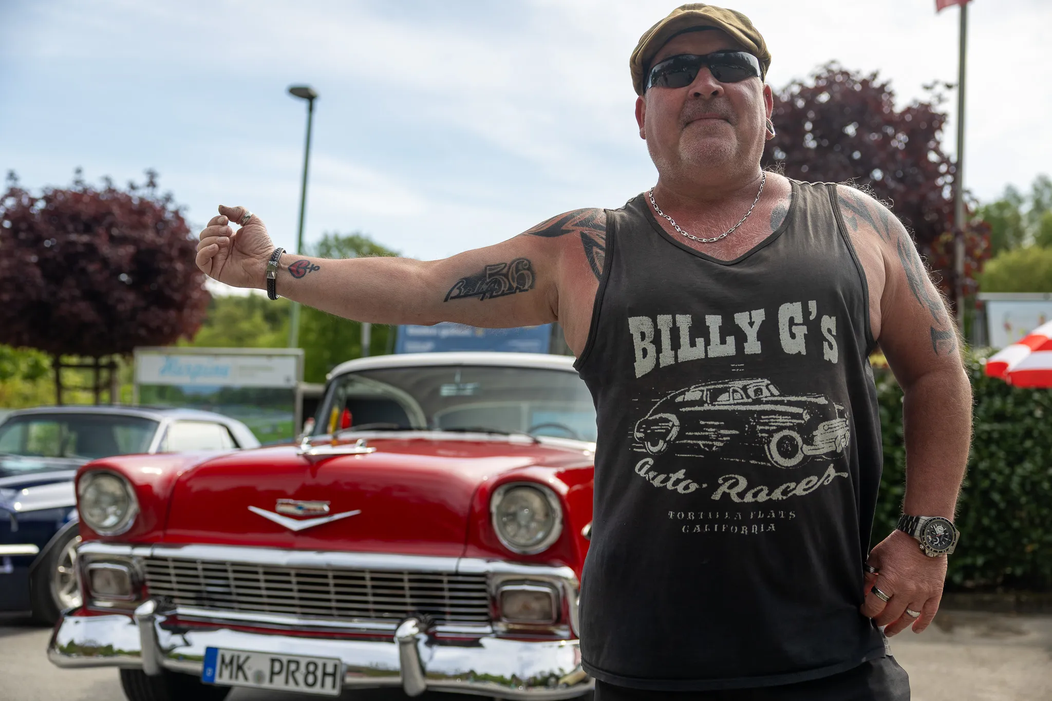 Mann mit Tätowierungen und Cap vor rotem Oldtimer, vermutlich auf einem Oldtimertreffen.