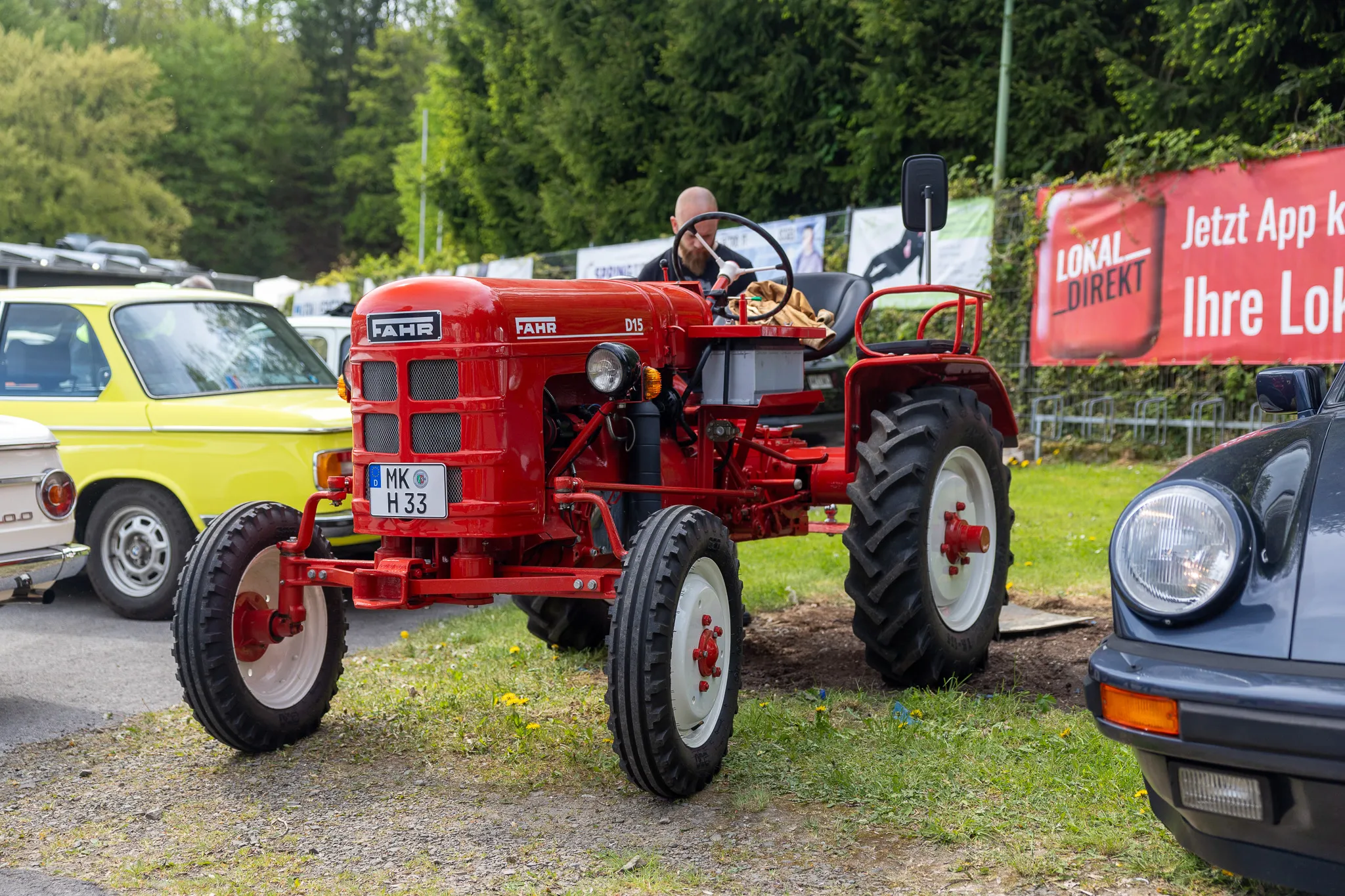 Roter Oldtimer-Traktor der Marke Fahr steht auf einer Veranstaltung zwischen anderen Fahrzeugen, darunter ein gelber Kleinwagen und ein schwarzer Sportwagen.