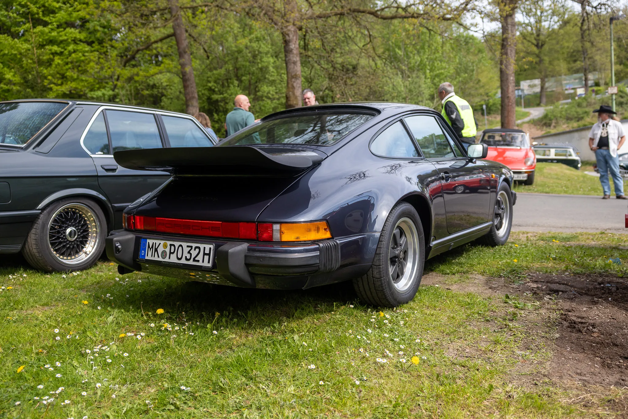 Dunkelblauer Porsche 911 mit Heckspoiler steht auf einer Wiese neben einem grauen Oldtimer. Im Hintergrund sind weitere Fahrzeuge und Personen zu sehen.