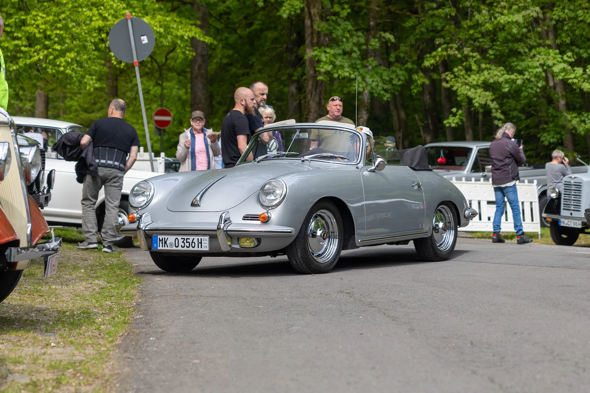 Silberner Porsche 356 Speedster fährt auf einer Straße vor einer Gruppe von Zuschauern bei einer Oldtimerausstellung.