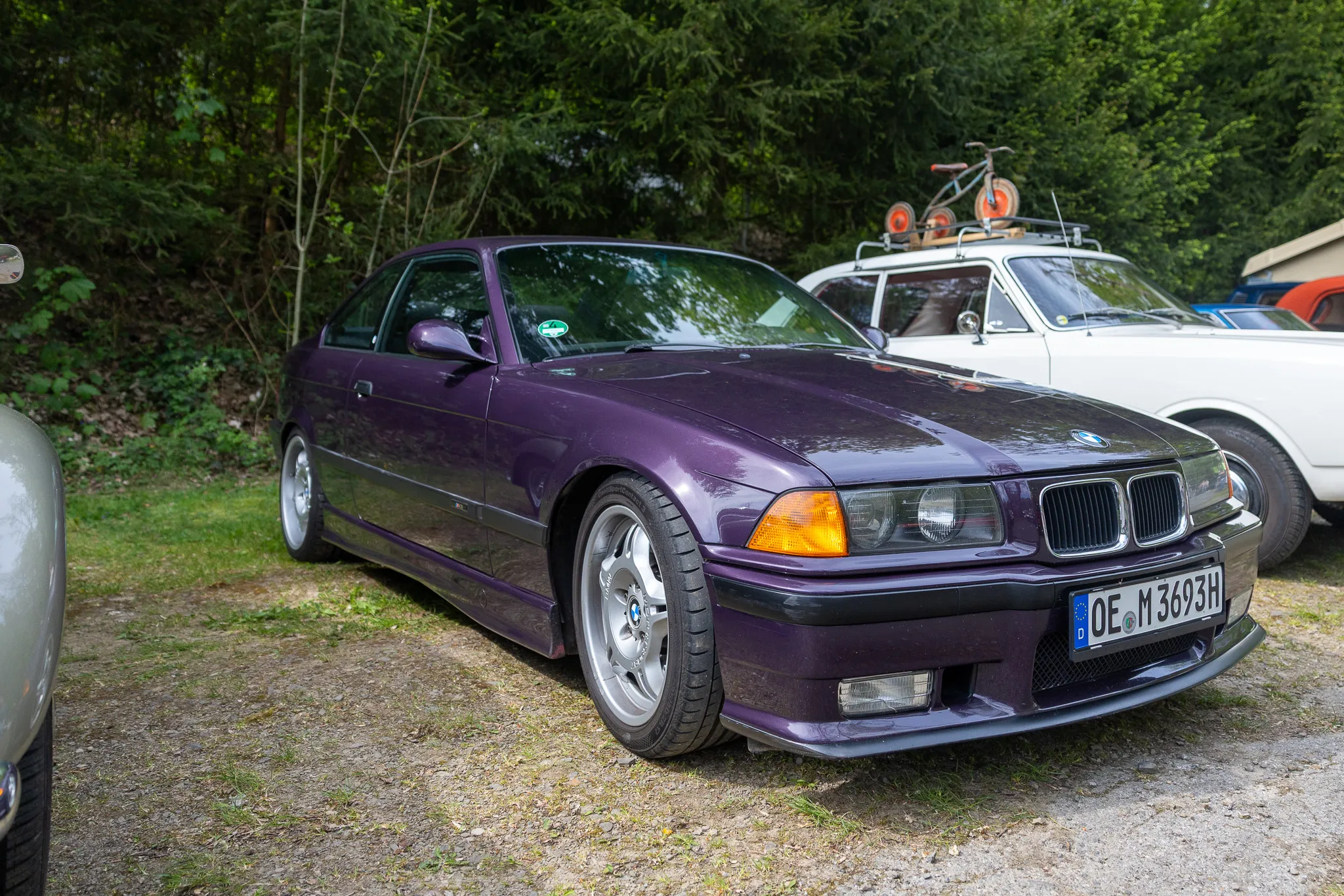 Dunkellila BMW der E36-Reihe steht auf einem Kiesplatz vor Bäumen, das Kennzeichen ist lesbar.