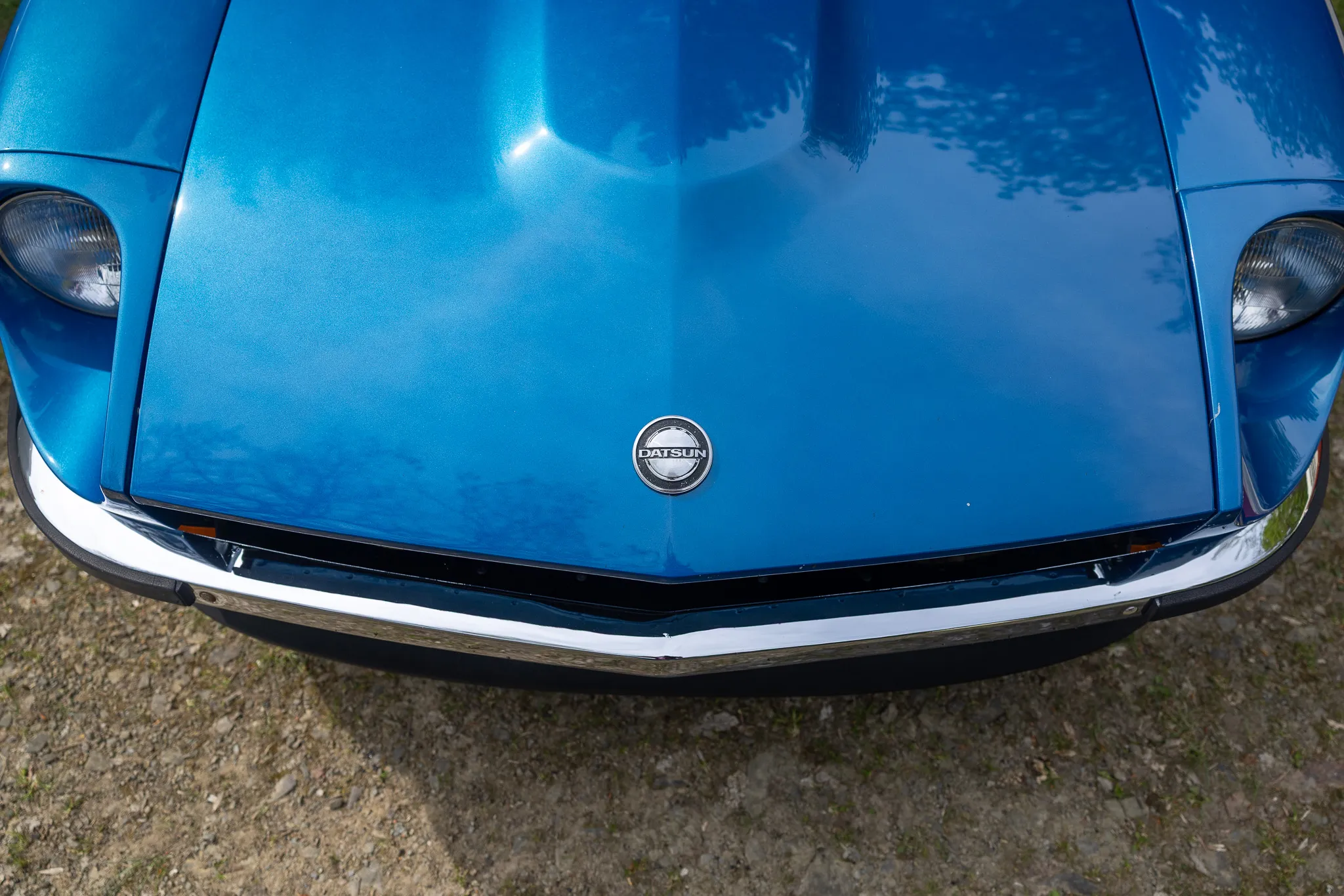 Nahaufnahme der blauen Motorhaube eines klassischen Datsun Sportwagens mit Chromstoßstange und zentralem Logo.