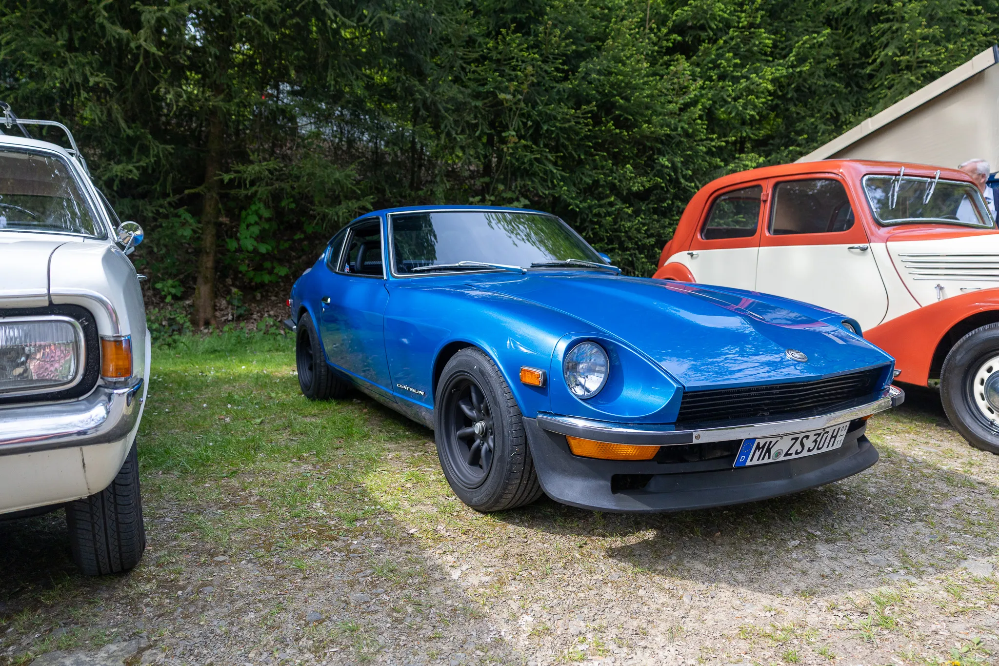 Blauer Datsun 240Z Sportwagen steht im Vordergrund eines Oldtimertreffens, umgeben von weiteren Fahrzeugen und Bäumen.