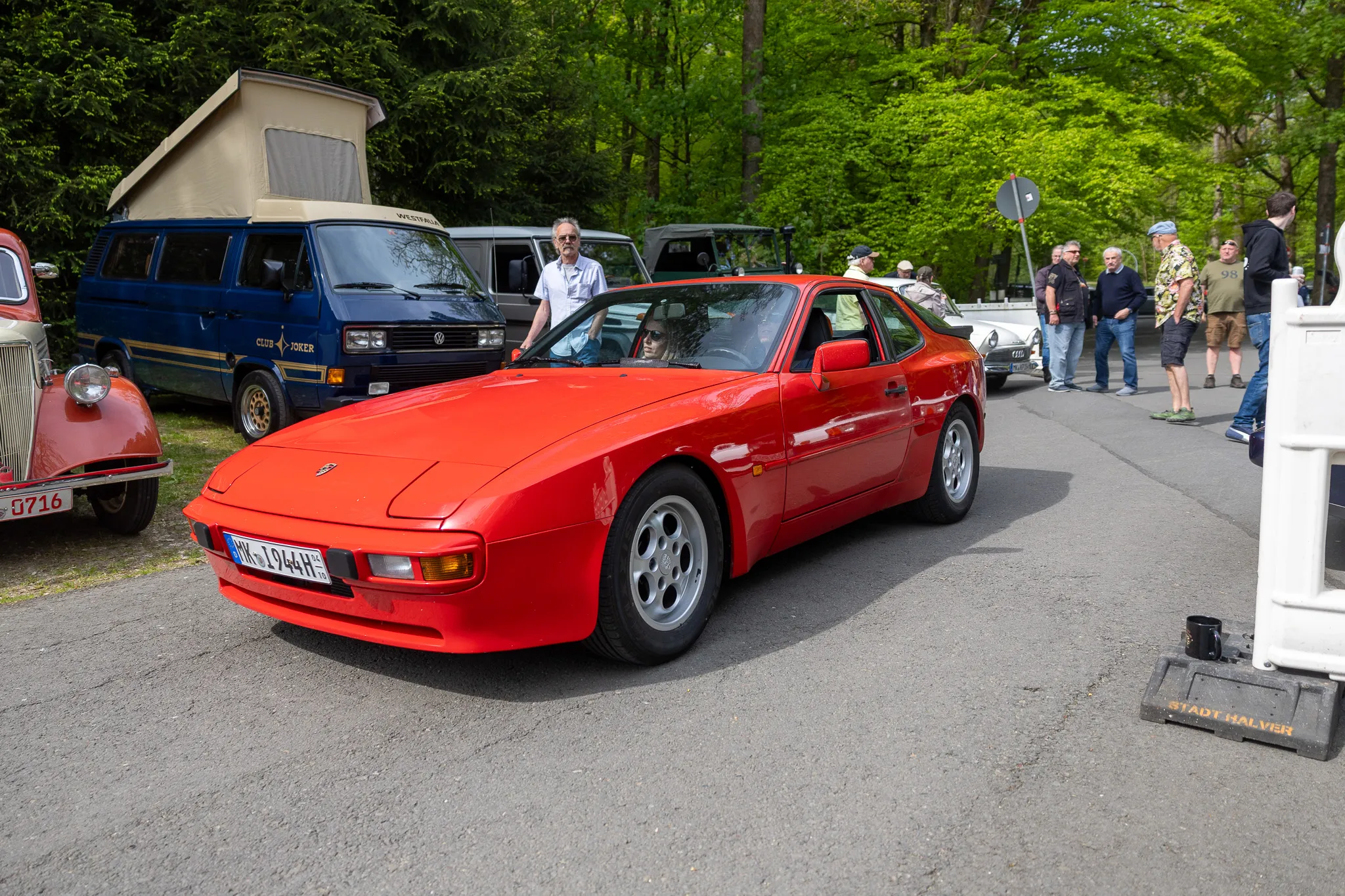 Roter Porsche 944 steht im Mittelpunkt eines Oldtimertreffens mit weiteren Fahrzeugen und Besuchern im Hintergrund.