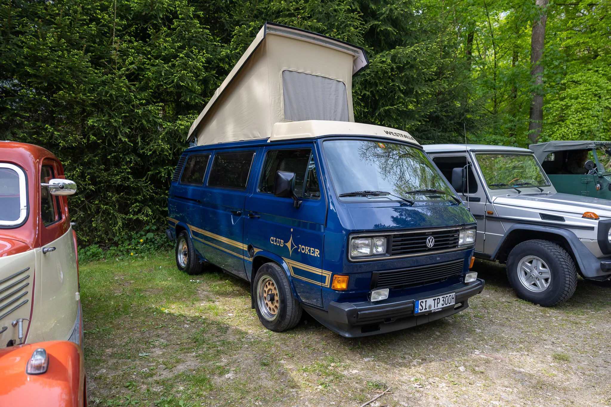 Blauer VW Westfalia Camper mit aufklappbarem Dach steht auf einem Kiesplatz vor Bäumen. Im Hintergrund ein gelber Oldtimer und ein grauer Geländewagen.