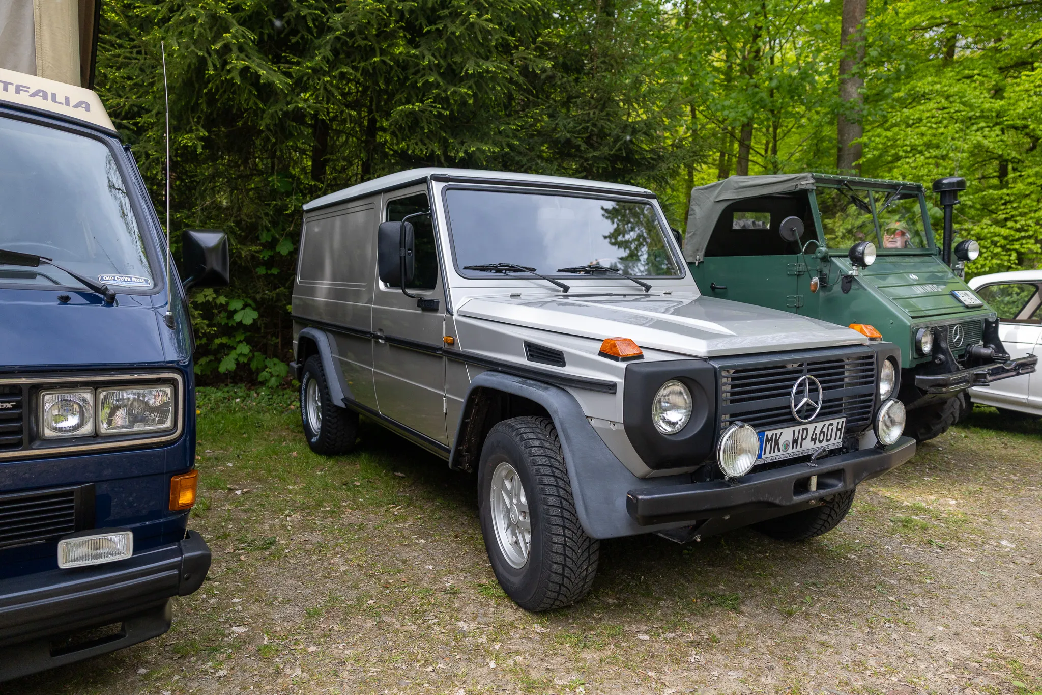 Grauer Mercedes-Benz G-Klasse Geländewagen steht neben einem blauen Transporter und einem weiteren Geländewagen im Wald.
