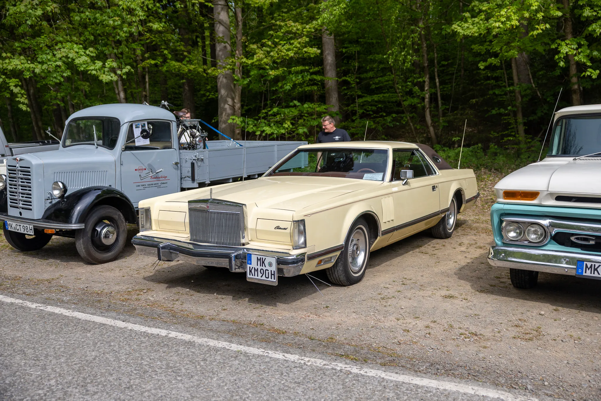 Beigefarbener Lincoln Continental Cabriolet steht im Vordergrund eines Oldtimertreffens, flankiert von einem weißen Lastwagen und einem blauen Pick-up.