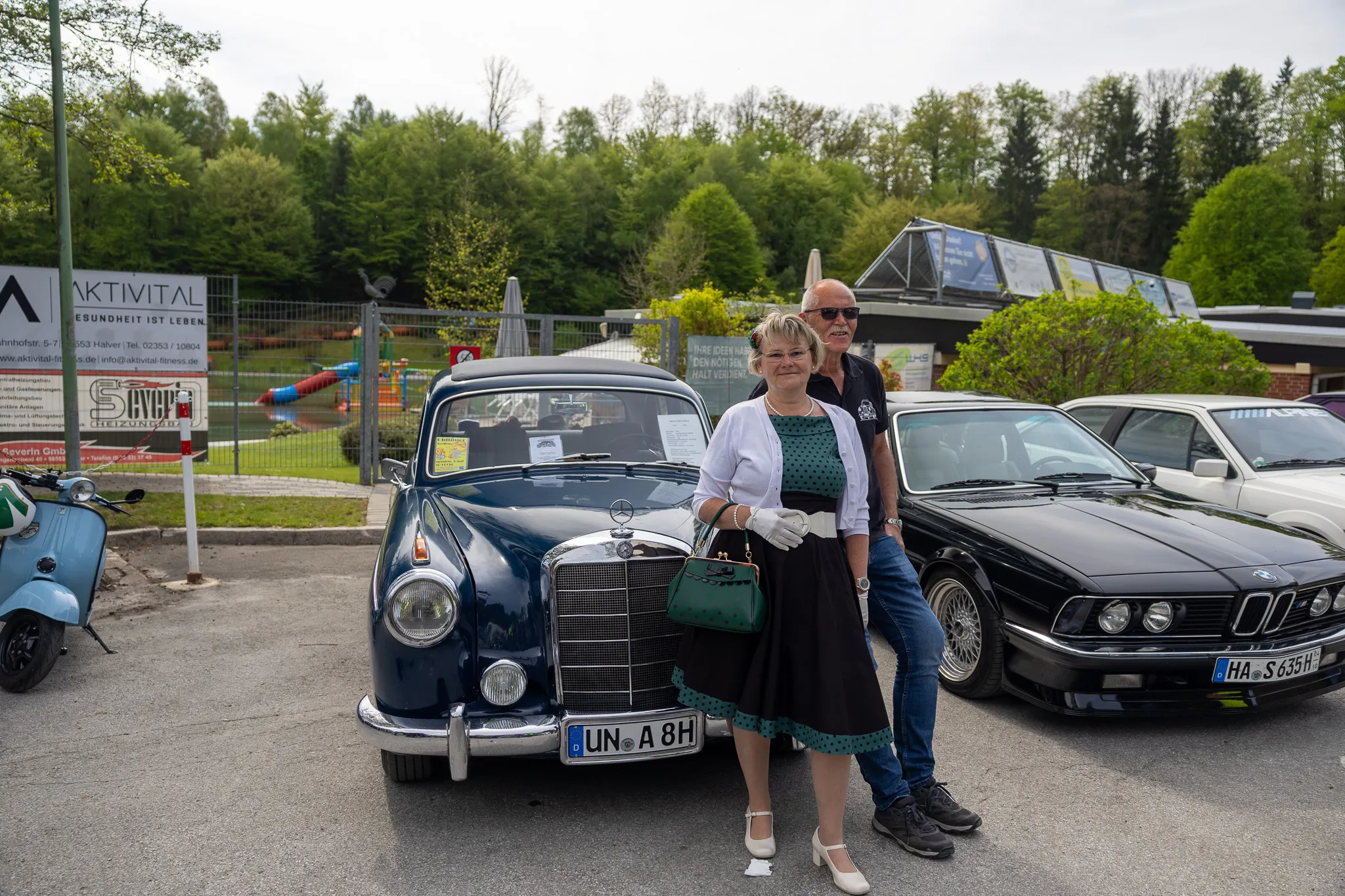 Ein älteres Paar steht lächelnd vor einem dunkelblauen Oldtimer mit geöffneter Motorhaube. Im Hintergrund sind weitere Oldtimer und ein Gebäude mit einem Glasdach zu sehen.