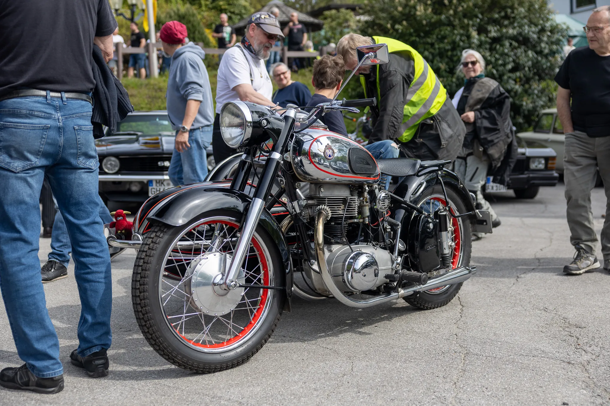 Detailaufnahme einer klassischen Motorradmaschine mit verchromten Teilen und rotem Streifen auf dem Reifen, umgeben von Besuchern eines Oldtimertreffens.