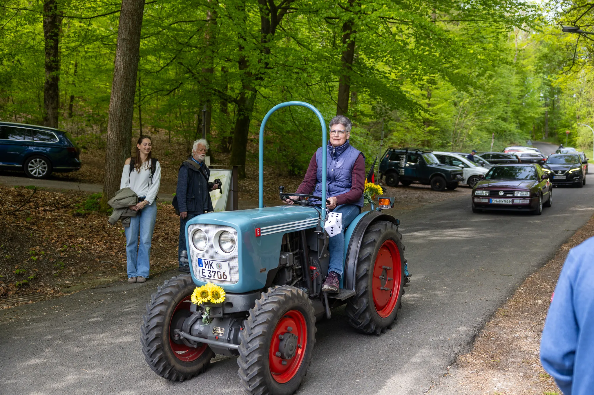 Eine Frau sitzt lächelnd auf einem restaurierten blauen Oldtimer-Traktor mit roten Rädern, während zwei Zuschauer zusehen. Im Hintergrund sind geparkte Autos und ein Wald zu erkennen.