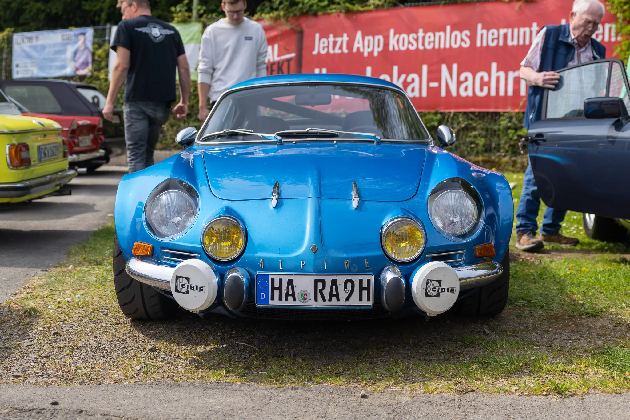 Blauer Alpine A110 Sportwagen mit zusätzlichen Scheinwerfern steht im Fokus eines Oldtimertreffens.