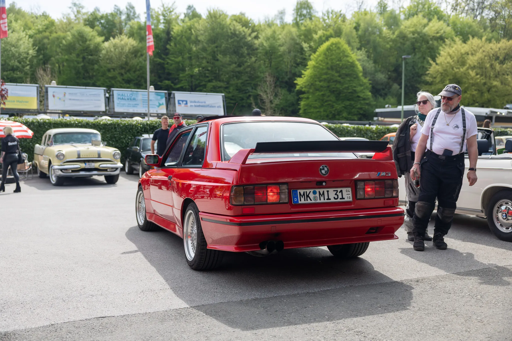 Roter BMW E30 Sport Edition steht auf einem Parkplatz, während ein Mann mit Kappe und Hosenträgern daran vorbeigeht. Im Hintergrund sind weitere Oldtimer und Zuschauer zu sehen.
