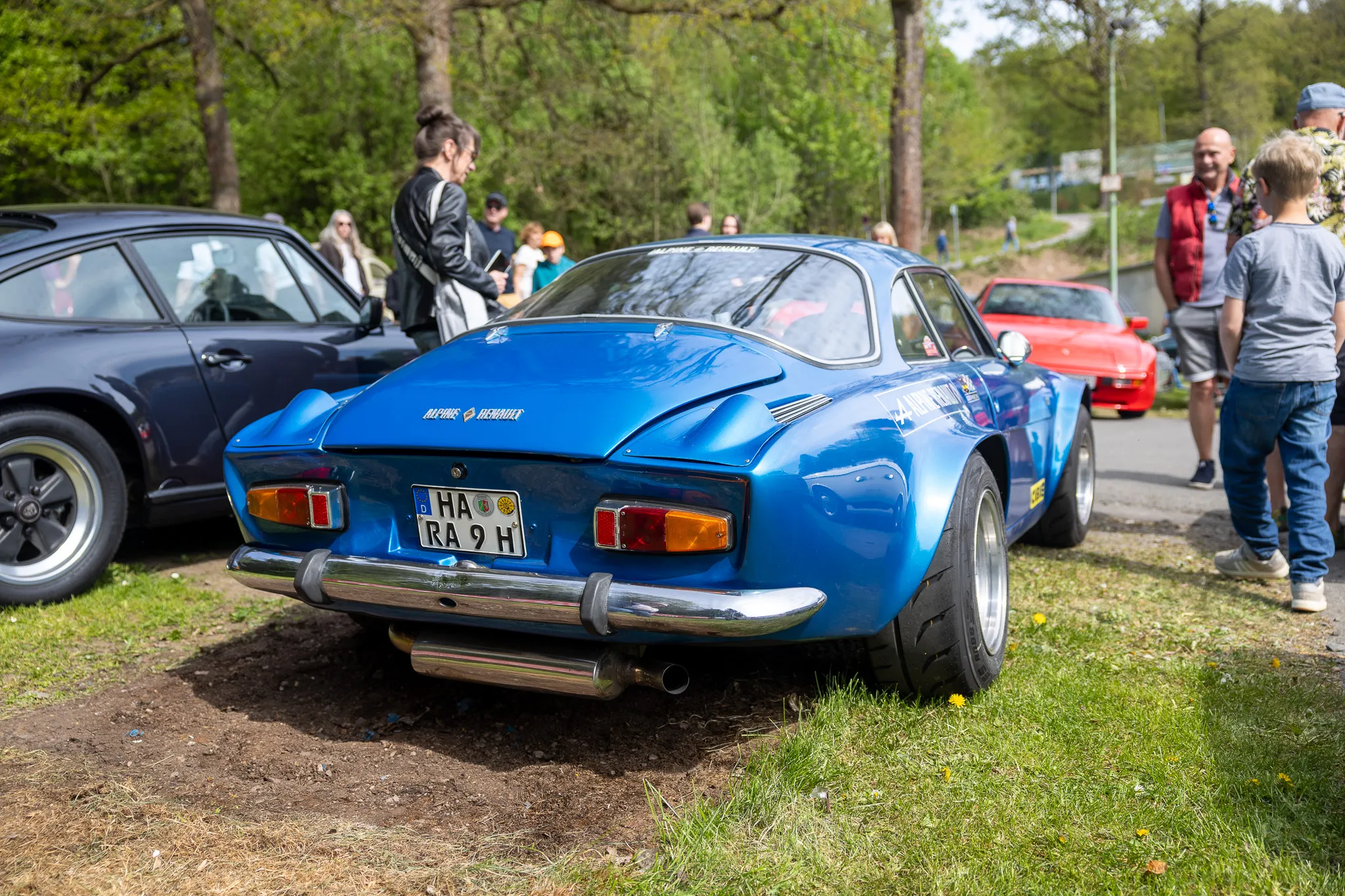 Blauer Alpine A110 steht im Mittelpunkt eines Oldtimertreffens, umgeben von weiteren Fahrzeugen und Besuchern.
