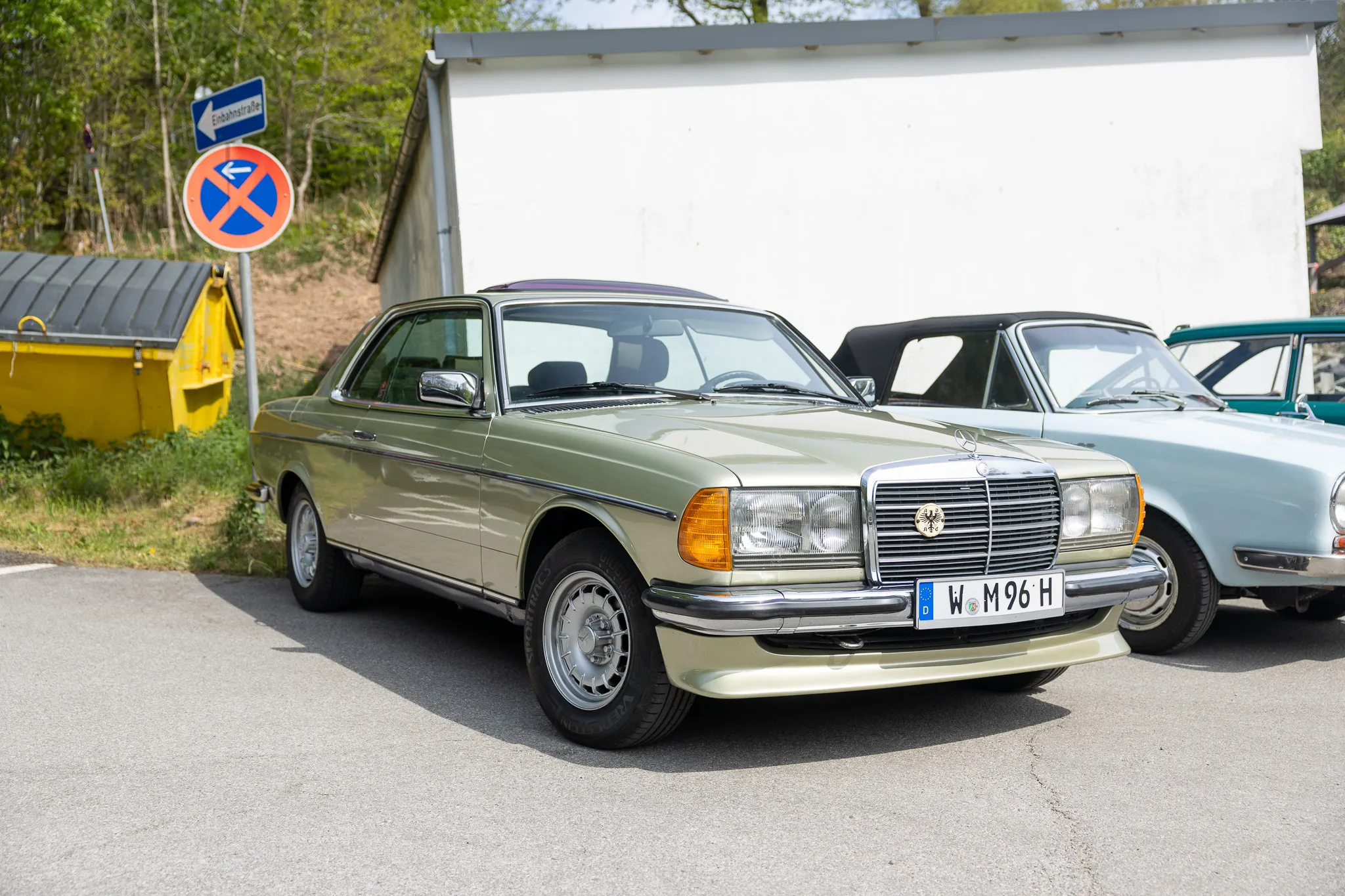 Hellgrüner Mercedes-Benz C123 Coupé steht auf einem Parkplatz vor Bäumen und weiteren Oldtimern. Im Hintergrund ein Verbotsschild und ein Straßenschild mit dem Hinweis „Einbahnstraße“.