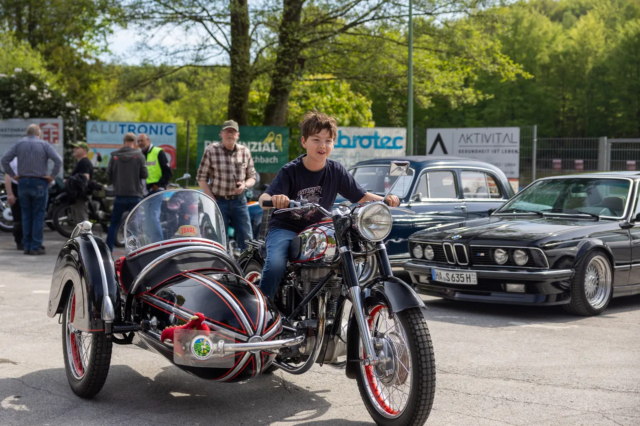 Ein Junge sitzt lächelnd auf einer klassischen Motorrad-Beiwagen-Kombination. Im Hintergrund sind weitere Oldtimer und Zuschauer zu sehen.
