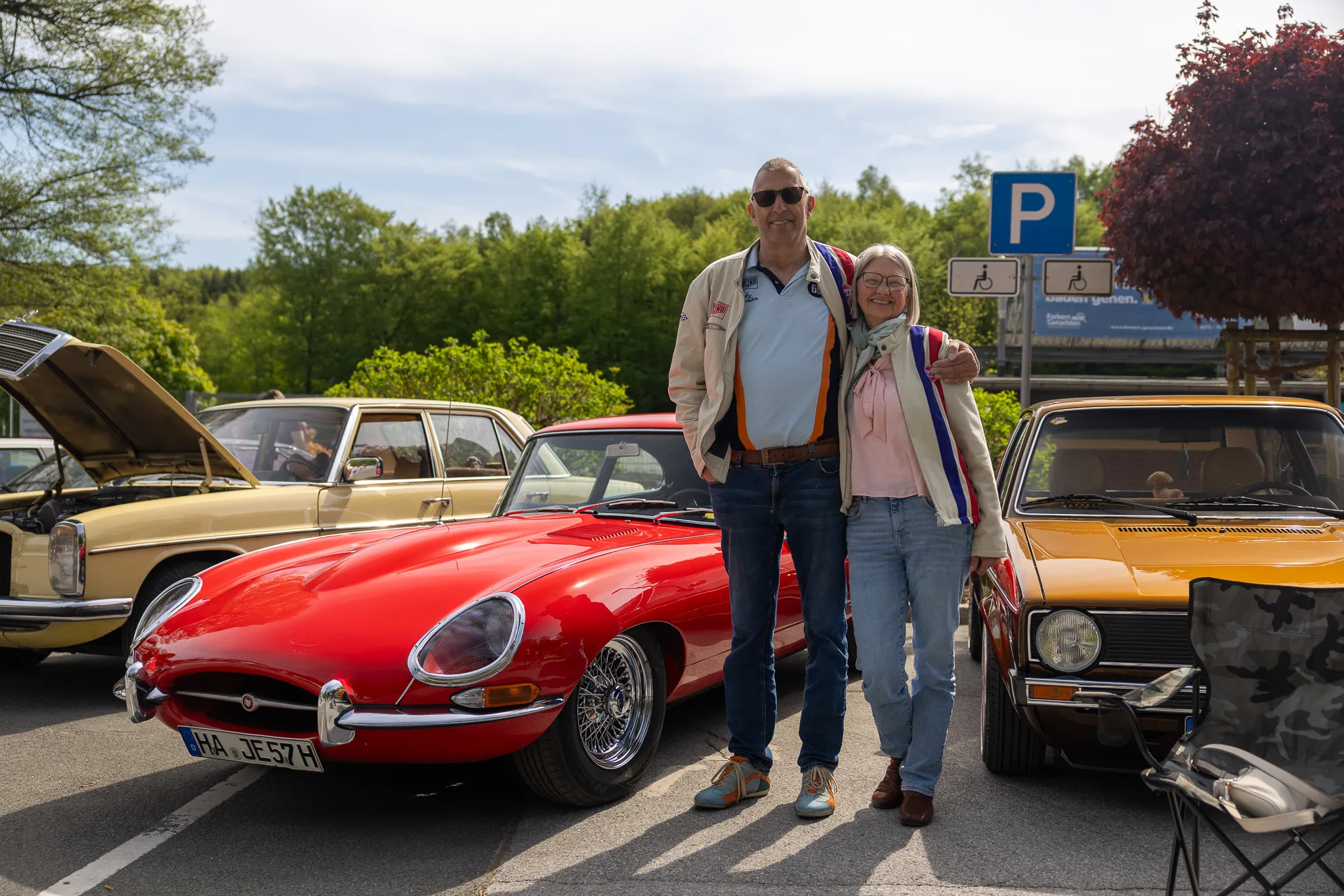 Ein Paar steht vor einem roten Oldtimer, umgeben von weiteren klassischen Fahrzeugen auf einem Oldtimertreffen.