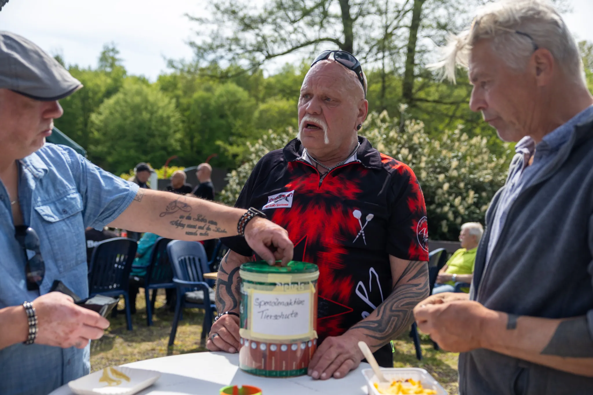 Ein Mann mit Schnurrbart schöpft mit einem Löffel aus einem großen Glas mit Spenden für den Tierschutzverein, während zwei weitere Männer zusehen.