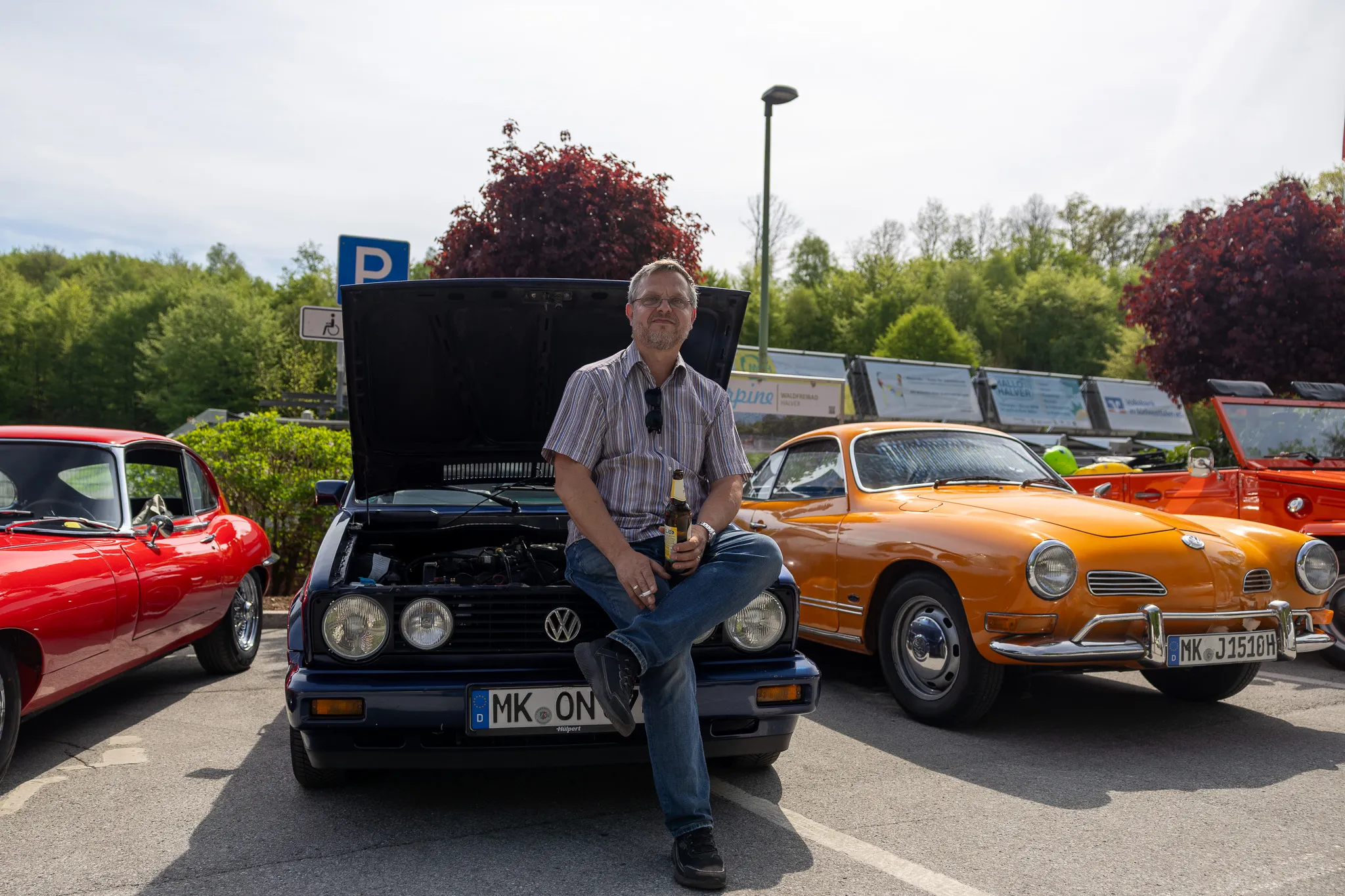 Ein Mann sitzt lächelnd mit einer Flasche Bier auf der Motorhaube eines schwarzen VW Scirocco. Im Hintergrund stehen weitere Oldtimer auf einem Parkplatz.