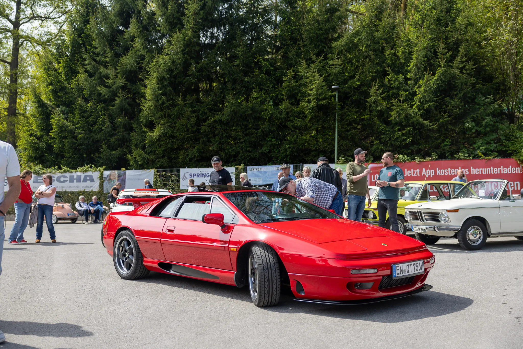 Roter Sportwagen steht im Mittelpunkt eines Oldtimertreffens mit weiteren Fahrzeugen und Besuchern im Hintergrund.