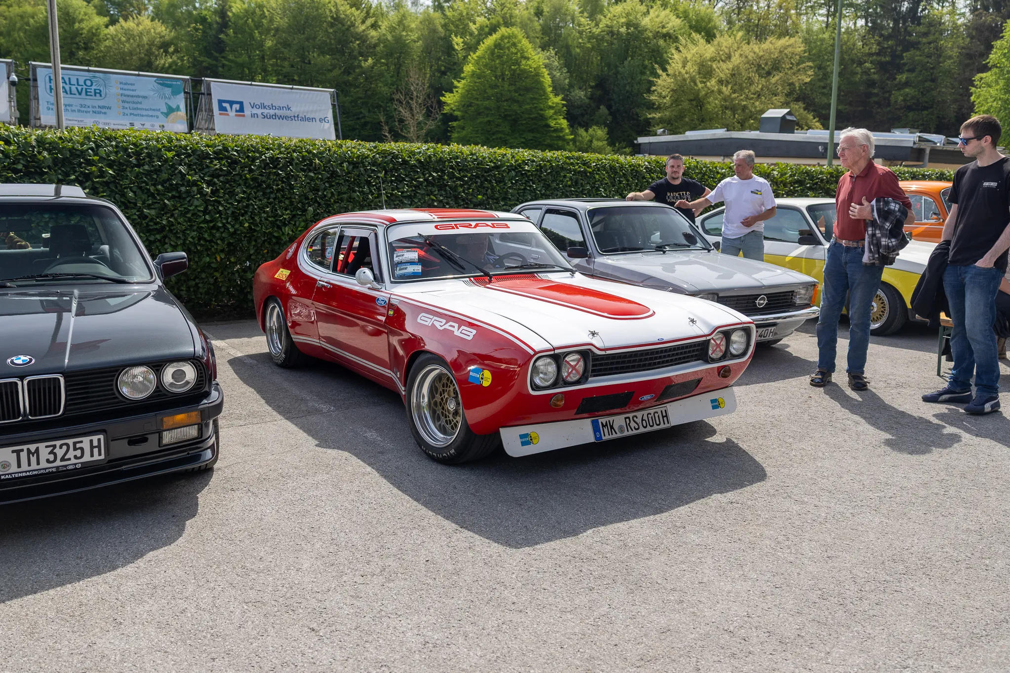 Roter Ford Capri mit weißen Streifen und dem Schriftzug „GRAB“ steht im Mittelpunkt eines Oldtimertreffens, umgeben von weiteren Fahrzeugen und Besuchern.