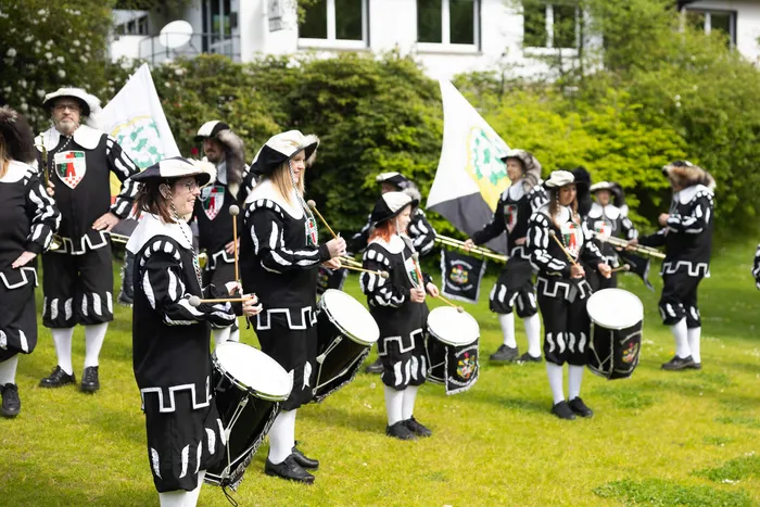 Eine Spielmannszug-Gruppe in historischen Uniformen mit Dreispitz und verzierten Jacken spielt auf Trommeln und Hörnern auf einer grünen Wiese.